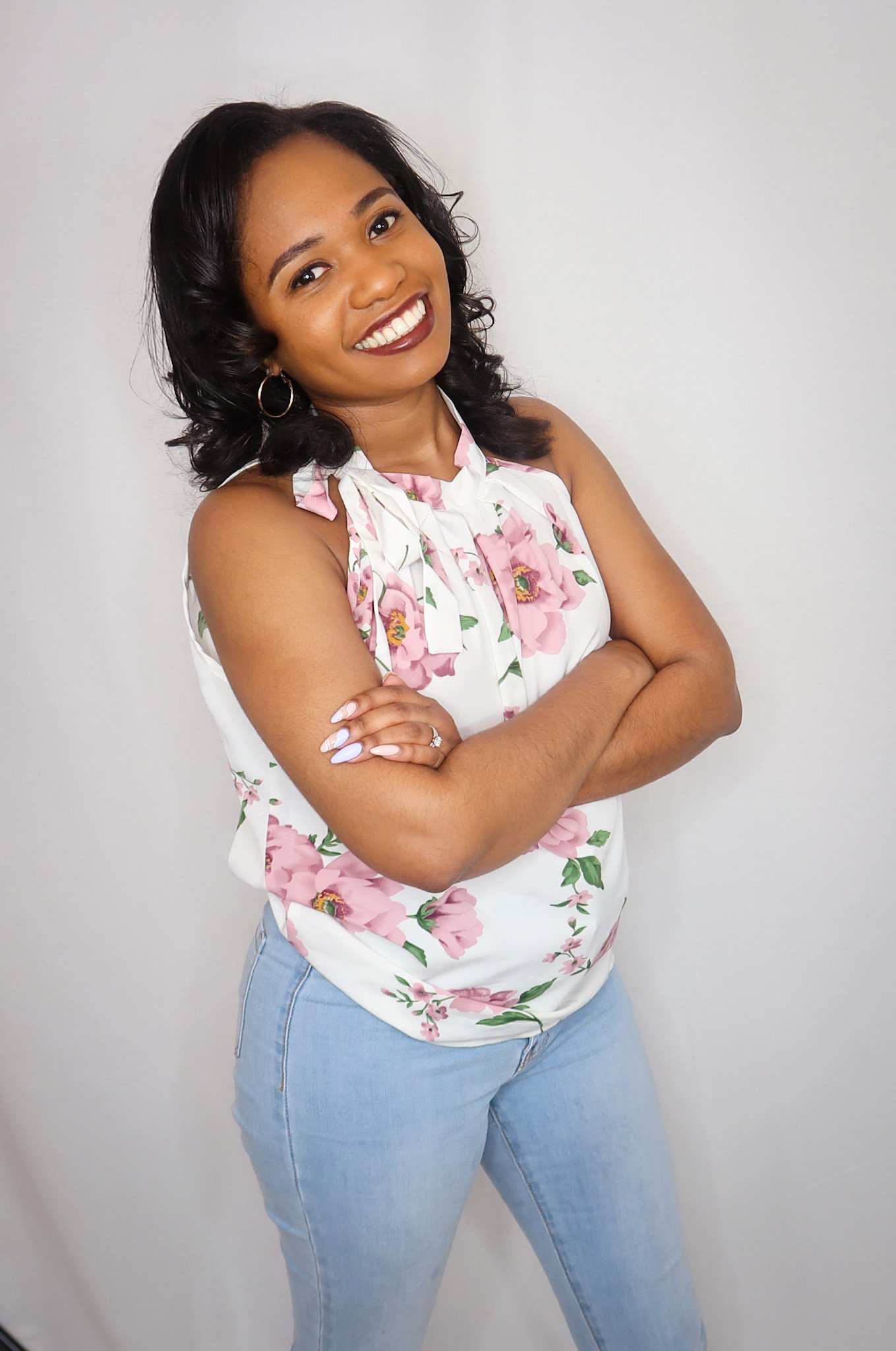 A woman with dark hair styled in loose curls, wearing a white sleeveless top with pink floral print and light blue jeans, standing with arms crossed and smiling at the camera against a plain white wall.