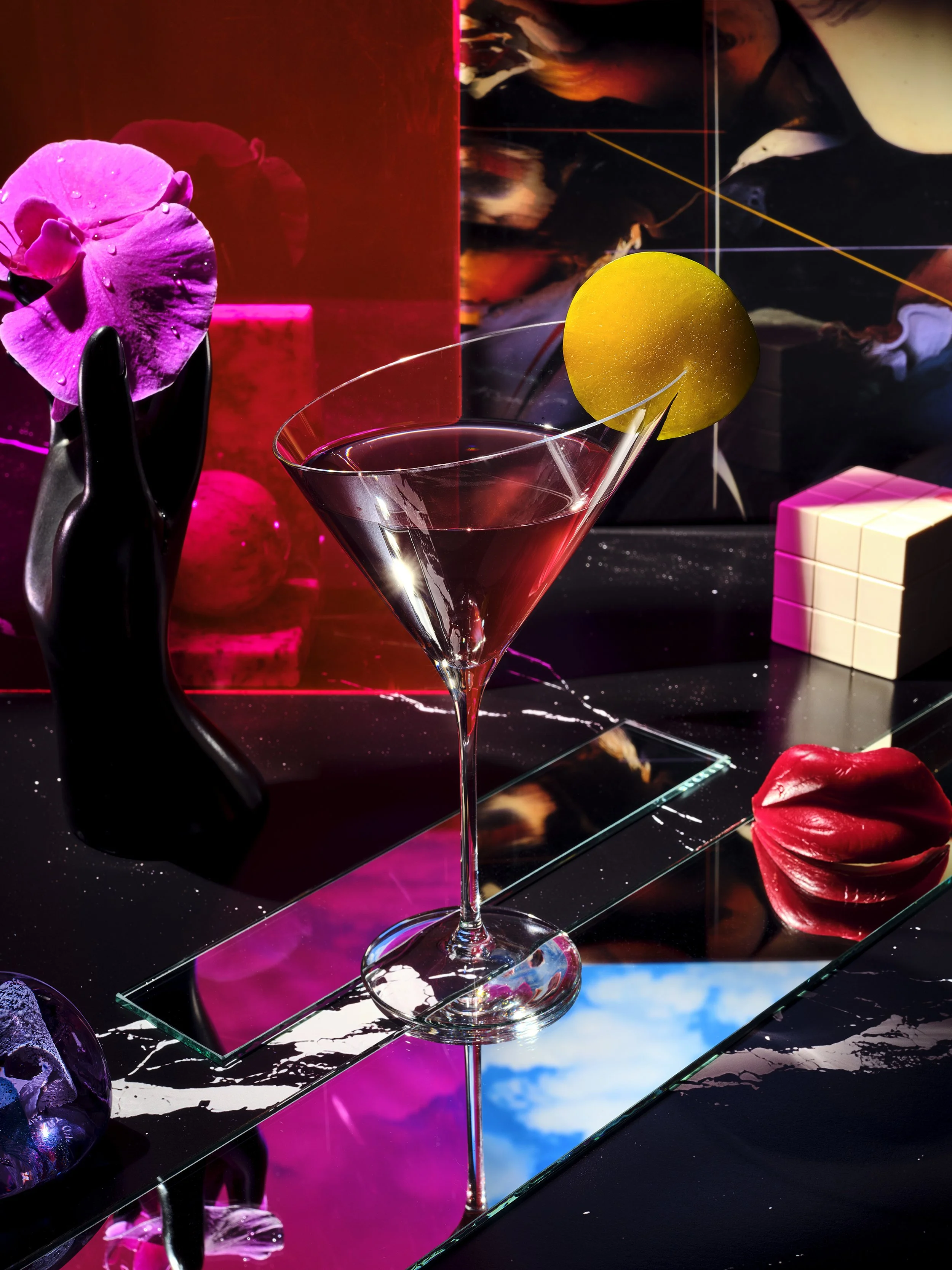 A cocktail glass with a lemon garnish surrounded by decorative objects including an orchid, a black abstract sculpture, a red lips sculpture, and colorful artwork in the background.