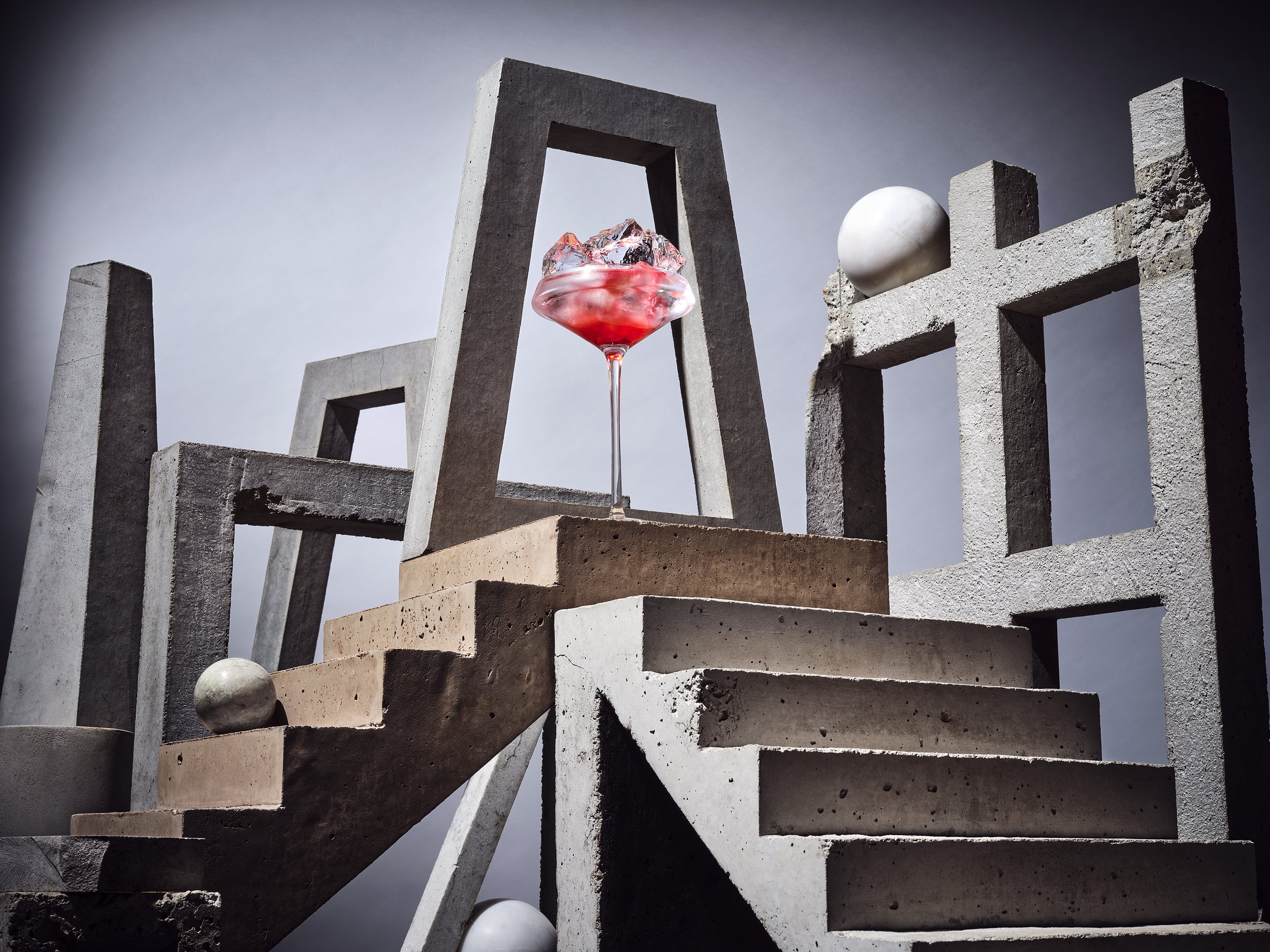 Abstract sculpture with concrete stairs, geometric blocks, and spheres, featuring a glass with a red drink and ice cubes on top of stairs.