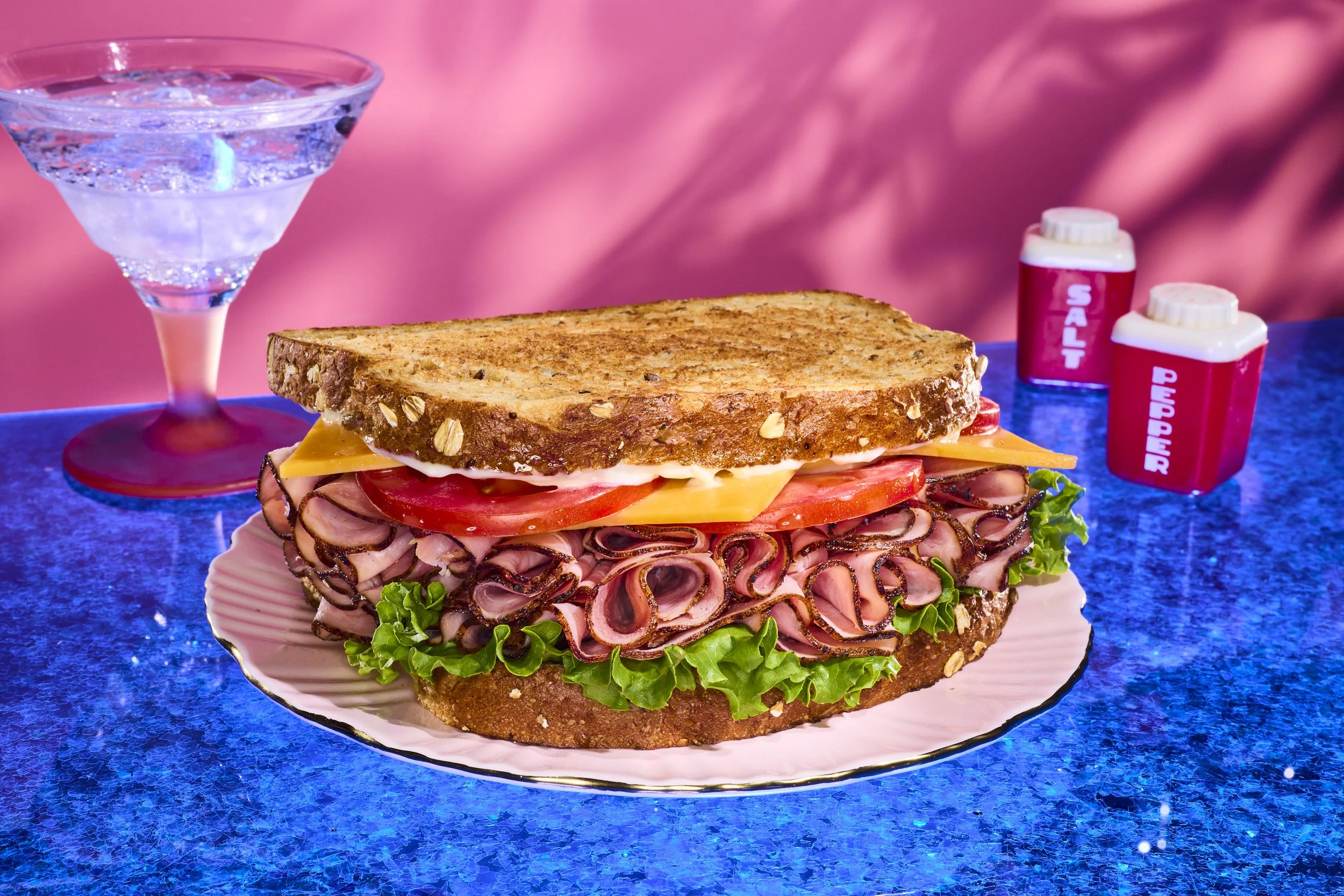 A large sandwich with roast beef, lettuce, tomato, cheese, served on toasted multigrain bread, on a decorative pink plate. There is a glass of sparkling water or soda, and two containers of salt and pepper shakers on the blue marble tabletop. The bac