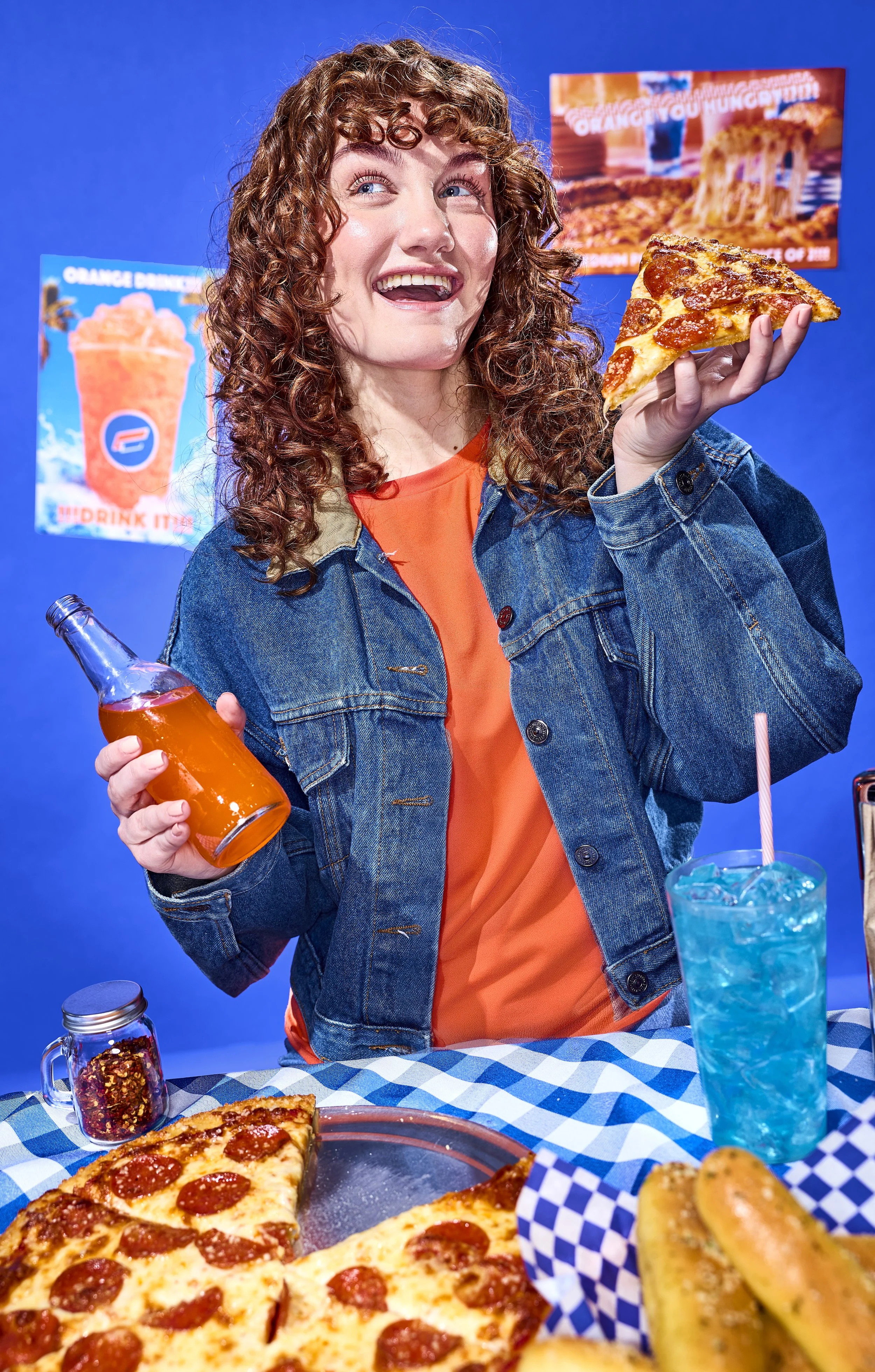 A woman with curly red hair wearing a denim jacket and orange shirt holding a slice of pepperoni pizza in one hand and a bottle of orange soda in the other, standing at a table with pizza slices, hot dog sandwiches, and a mason jar with red pepper fl