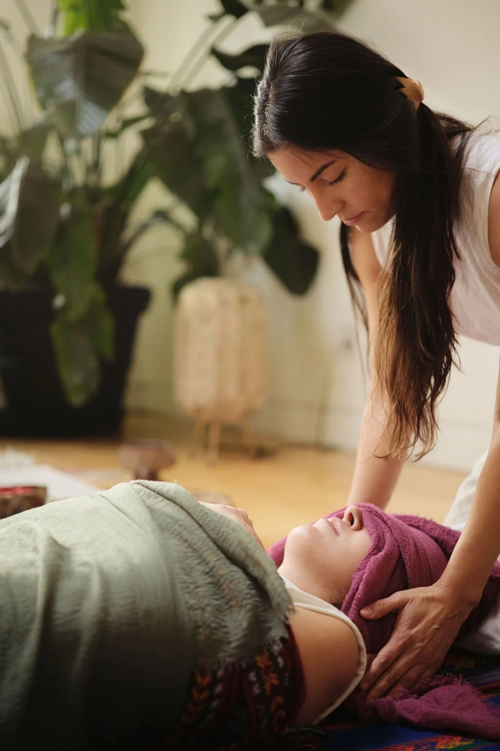 A woman giving a massage to a person lying down with a purple towel on their head. The woman is focused and gentle, in a cozy, well-lit room with plants and a wicker lamp in the background.