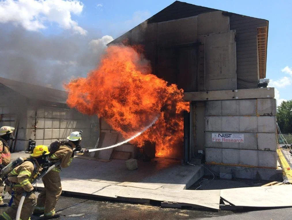 Three firefighters holding a hose and aiming water at flames coming out of the entrance to a warehouse-style building.