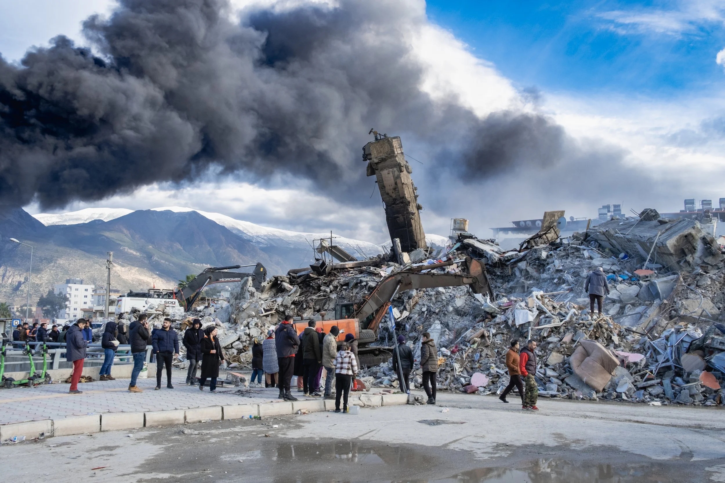Collapsed buildings with a crowd of men standing in the foreground, black smoke plume overhead, and mountains in the distance