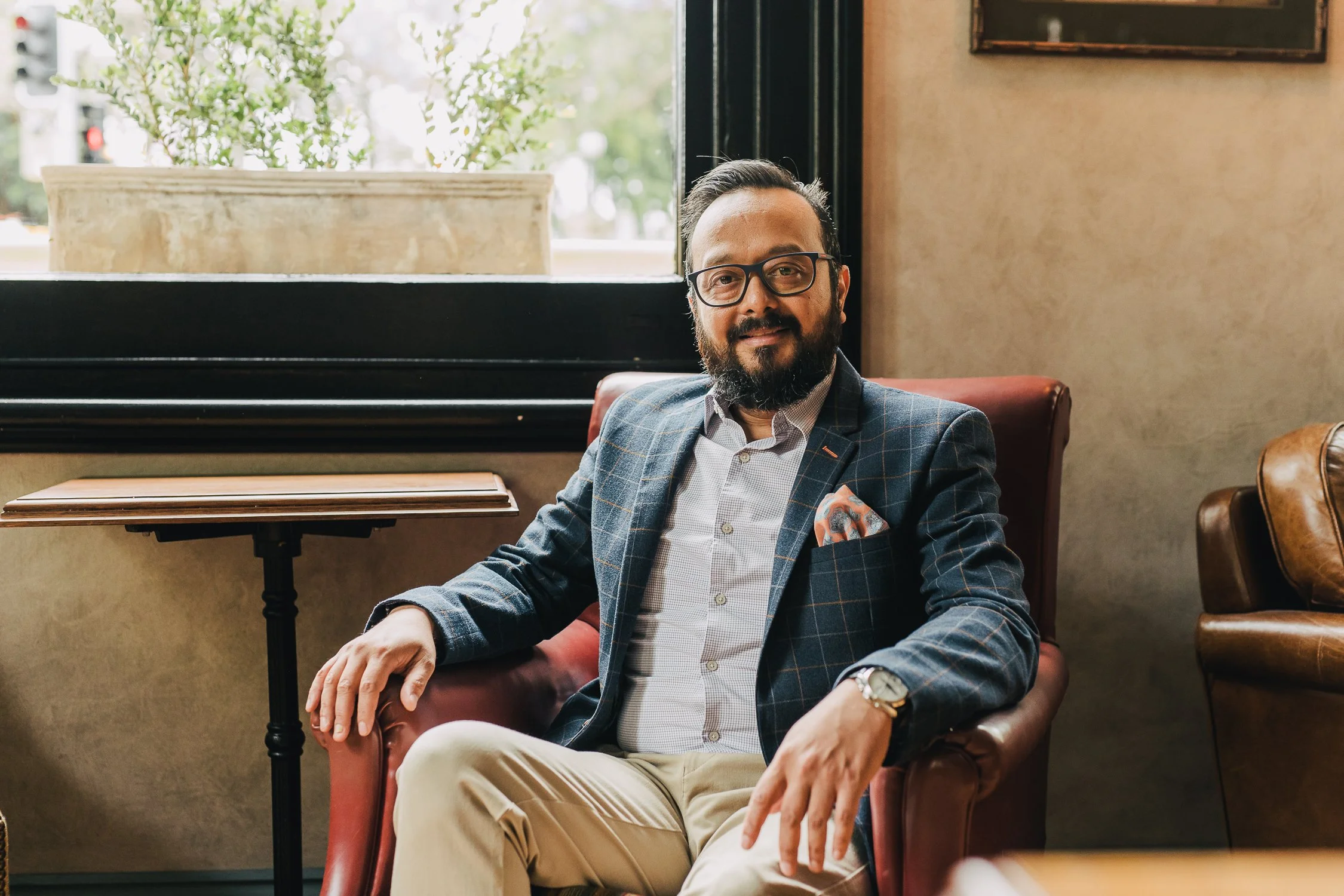 A man with glasses, a beard, and dark hair, dressed in a plaid blazer, light shirt, and beige pants, seated in a leather armchair inside a café or restaurant with a window and plant behind him.