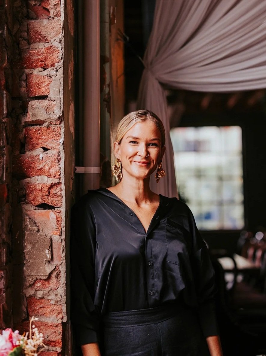 A woman with blonde hair, gold earrings, and a nose ring, smiling and standing indoors near a brick wall and a window with a curtain.