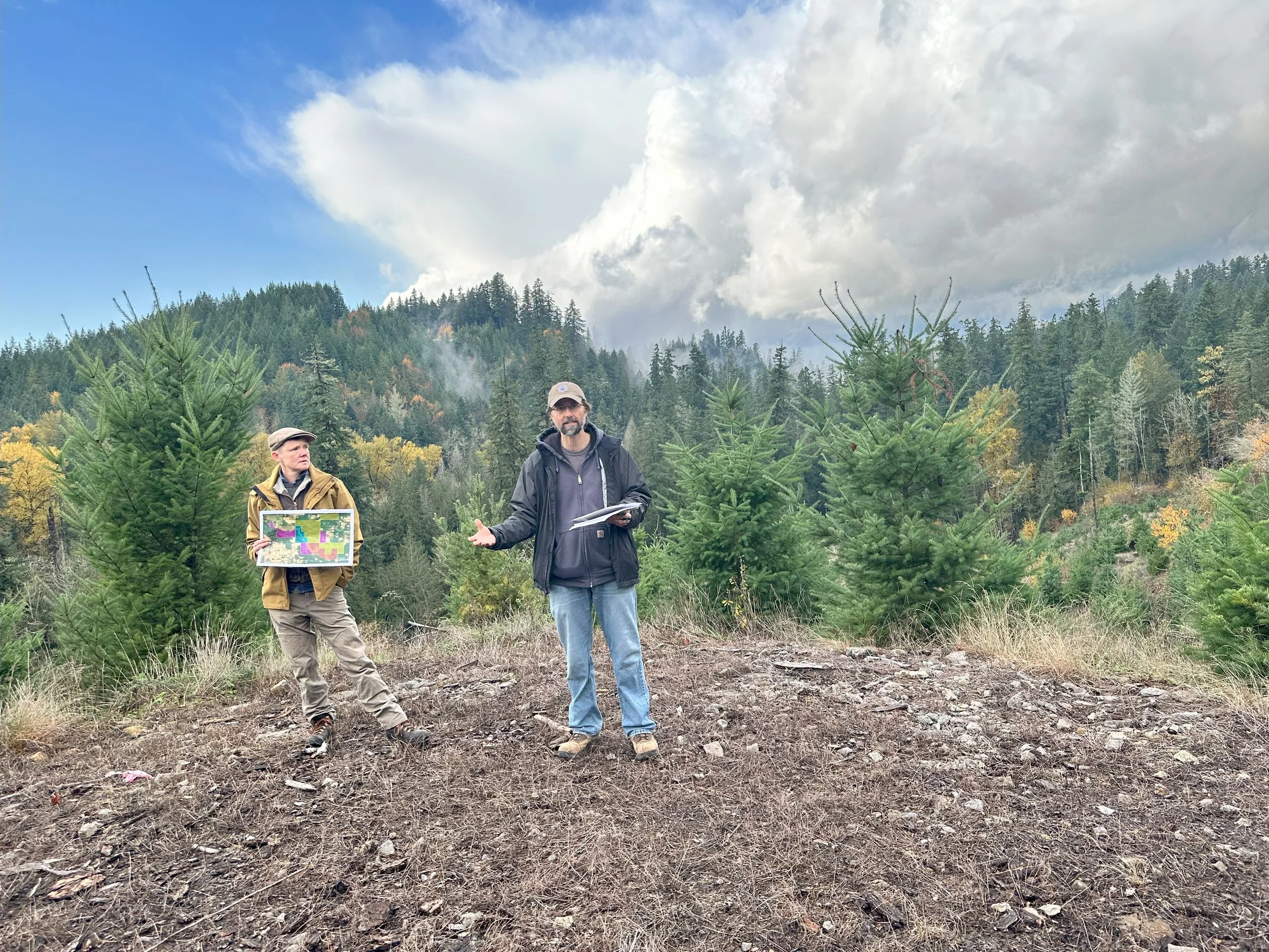 Two people stand presenting in front of a forested hillside. One is holding a map and the other is speaking.
