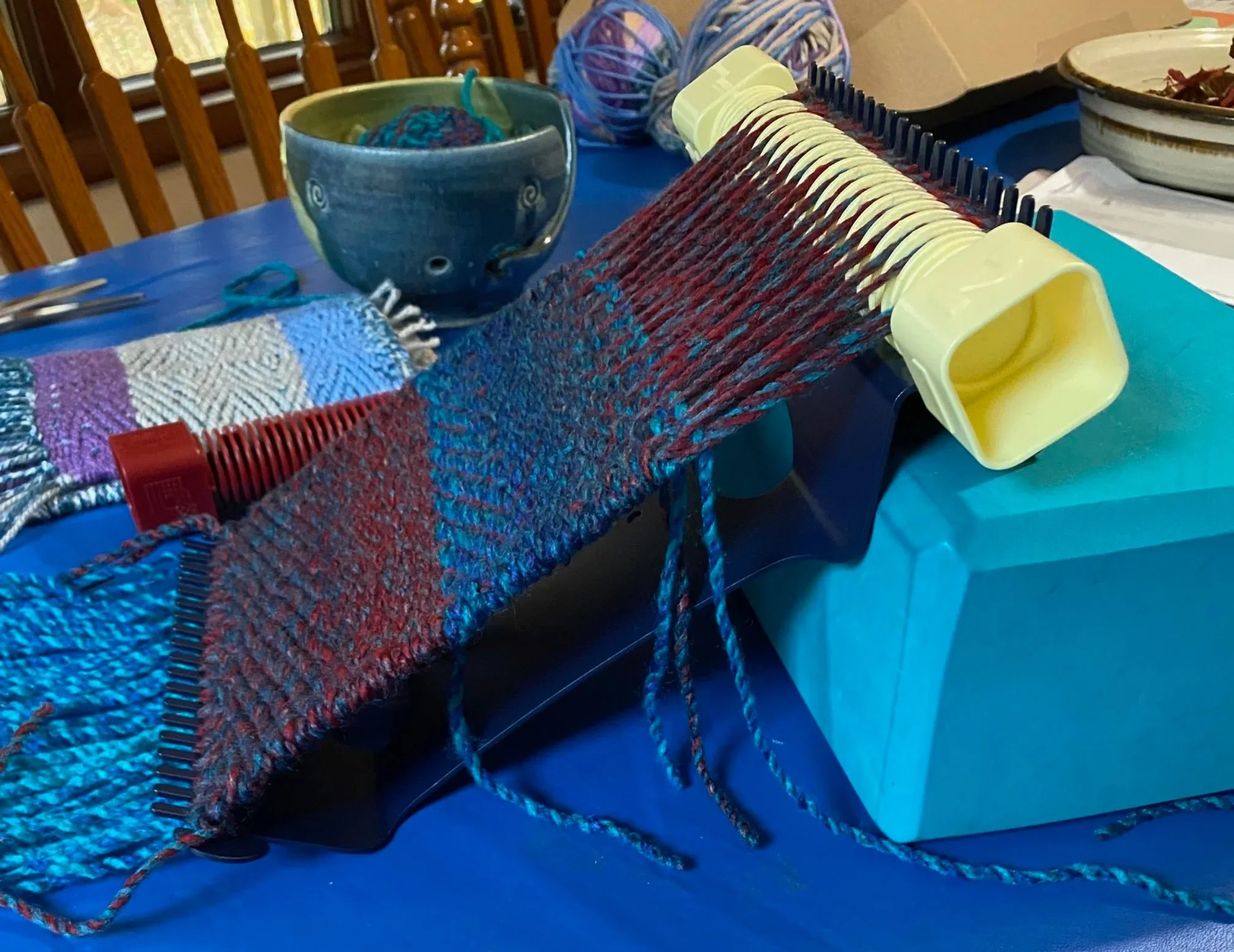 Colorful weaving in progress on a small handheld loom, with yarn and weaving tools on a table.