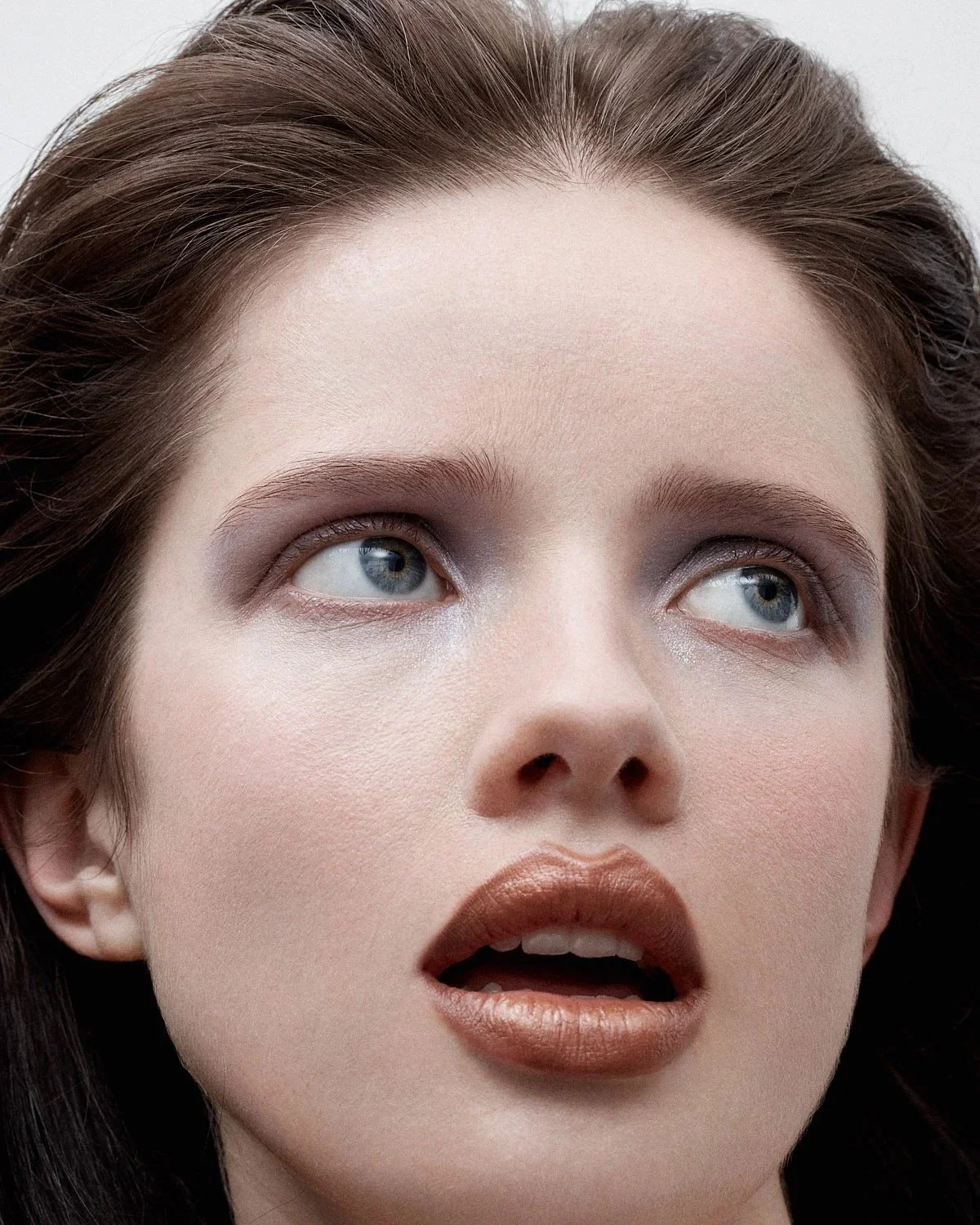 Close-up of a woman's face with makeup, blue eyes looking upward, lips slightly parted, and brown hair.