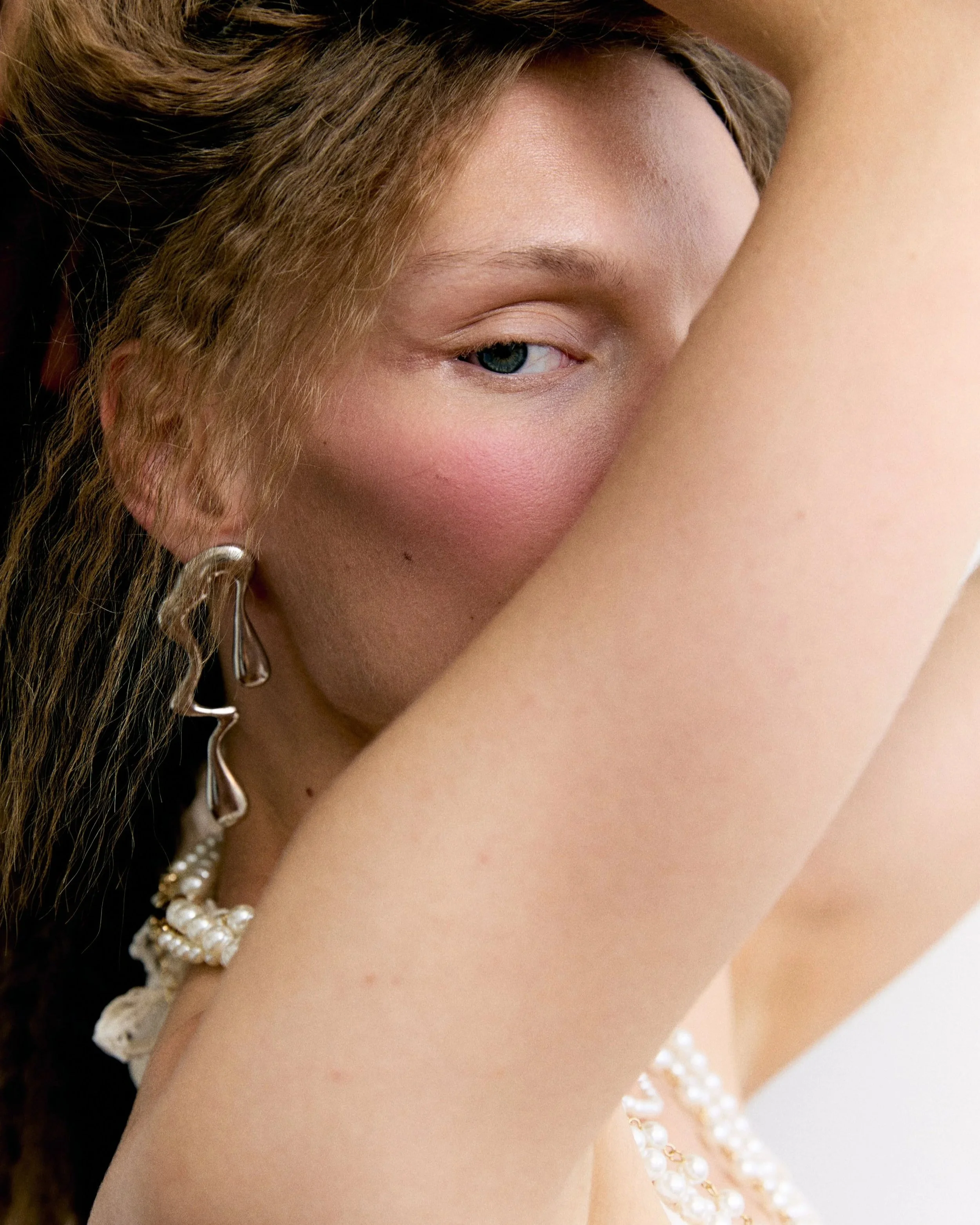 Close-up of a woman's face, showing her eye, nose, and part of her lips, with her arm covering part of her face. She has light skin, wavy brown hair, and wears pearl jewelry.