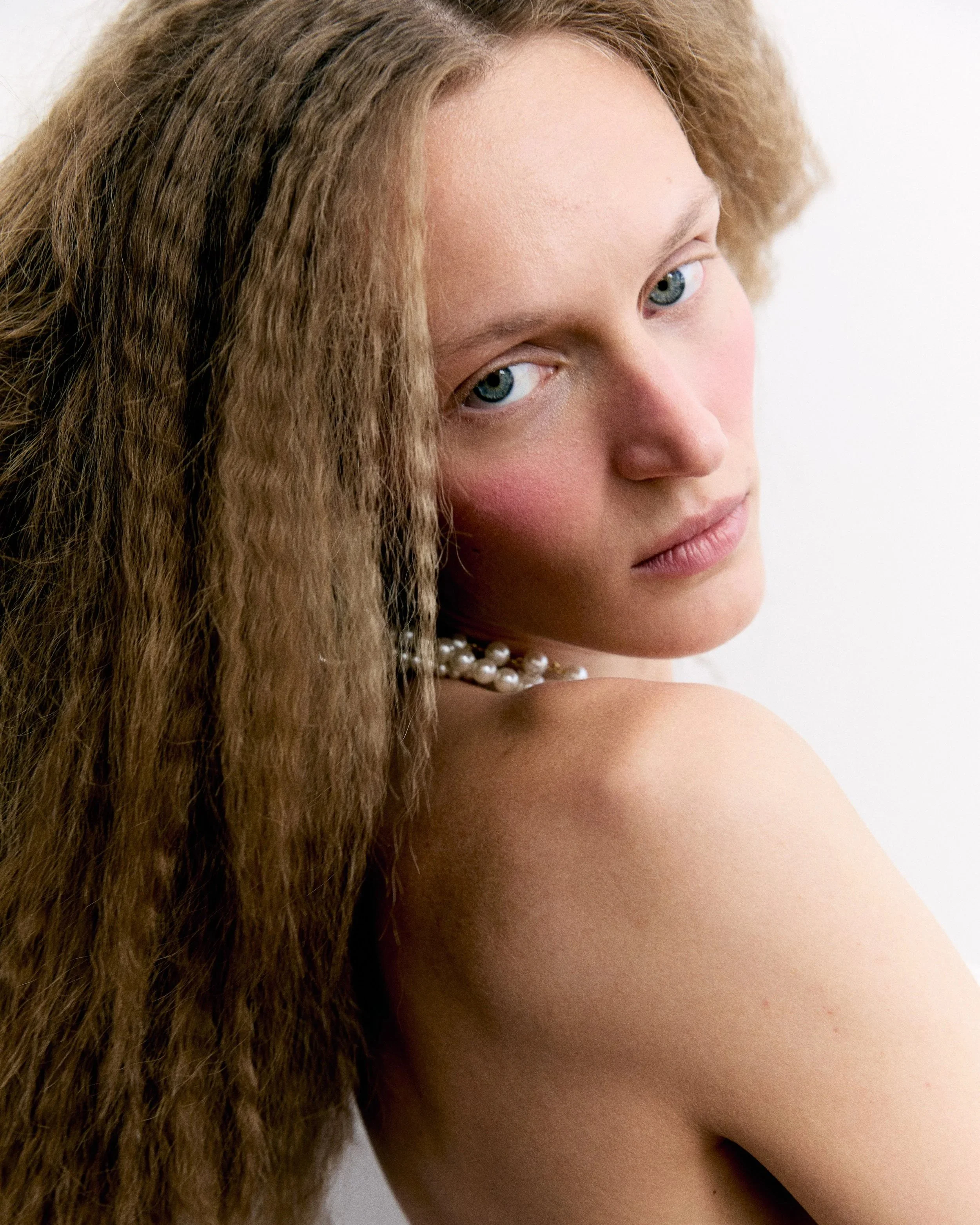 Close-up of a woman with long, curly brown hair, blue eyes, light pink lips, wearing a pearl necklace, and posing with her face turned slightly to the side against a white background.