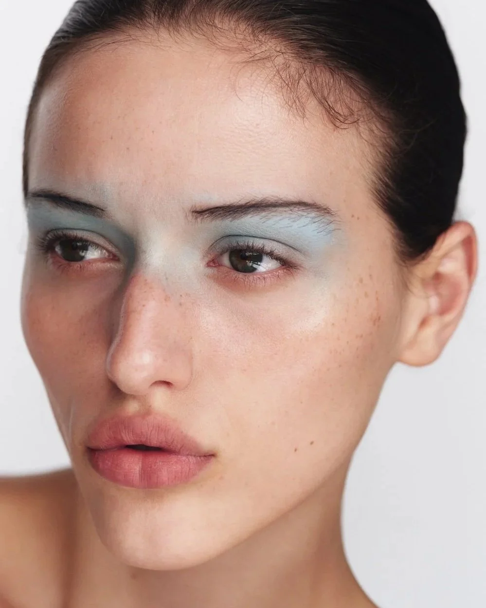 Close-up of a woman's face with minimal makeup, showcasing natural skin with freckles, light eyeshadow, and neutral lips.