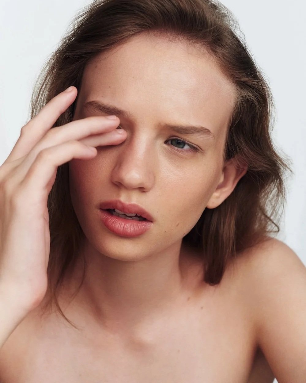 A young woman with brown hair touching her face and looking slightly confused or tired.
