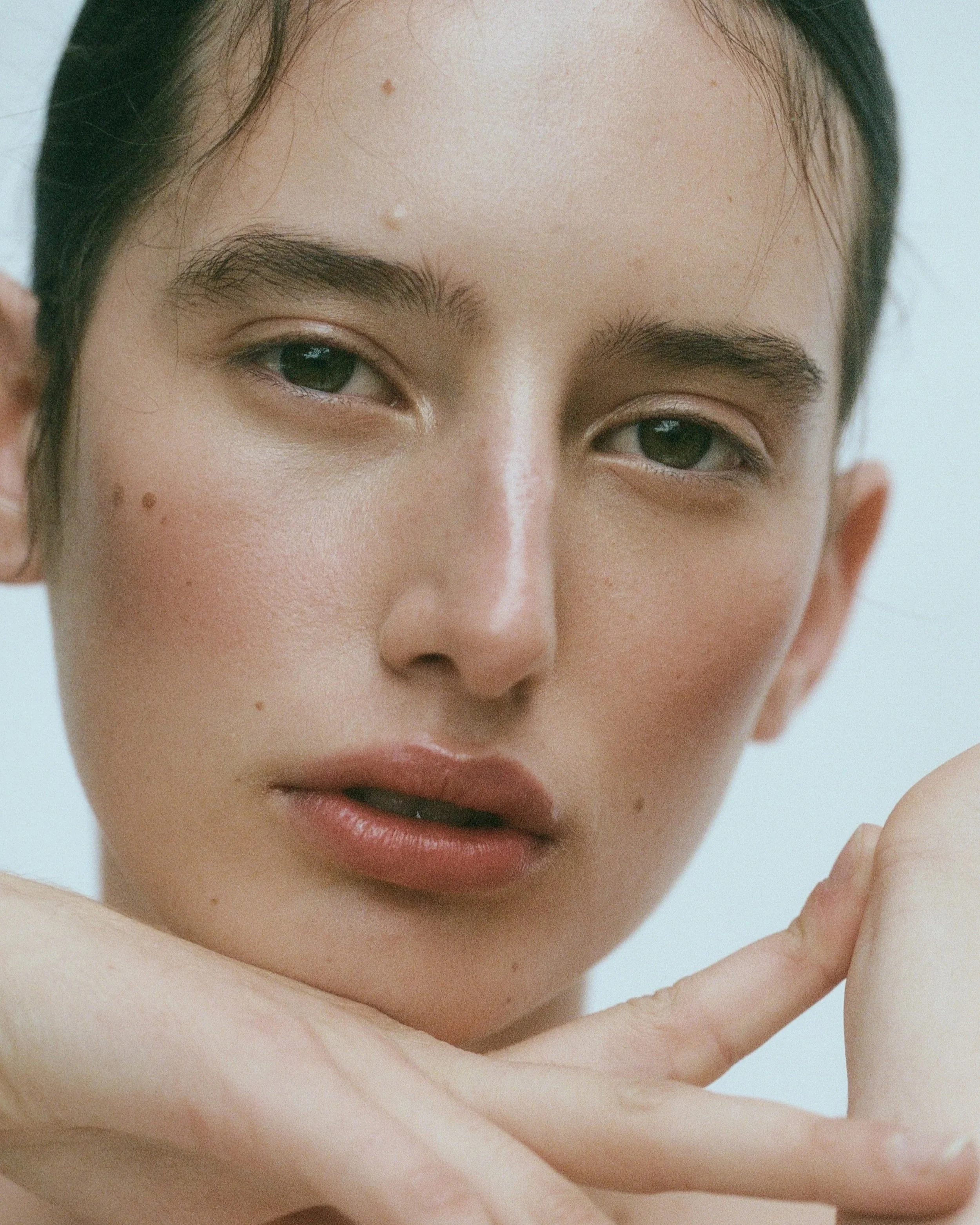 Close-up of a young woman with fair skin, dark hair, and green eyes, posing with her hand touching her face.