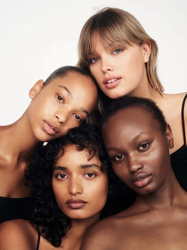 Group of four diverse women with different skin tones and hair styles posing together against a white background.