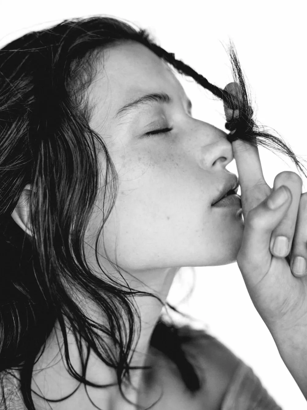 A black and white close-up portrait of a young woman with wavy hair, eyes closed, holding her finger to her lips in a shushing gesture.