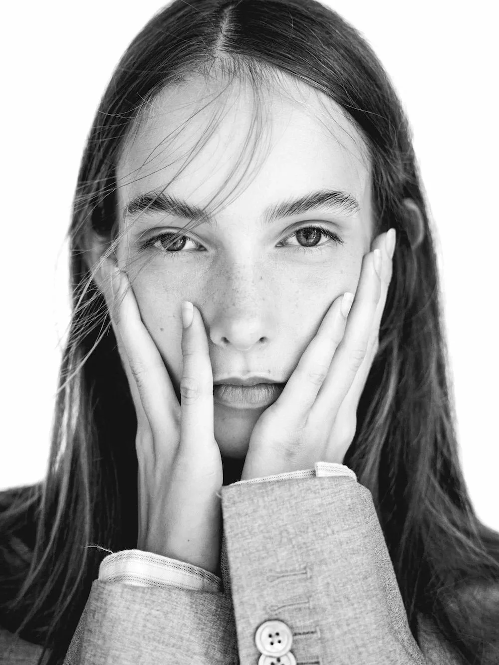 Black and white portrait of a young woman with long hair, holding her face with both hands, looking directly at the camera.