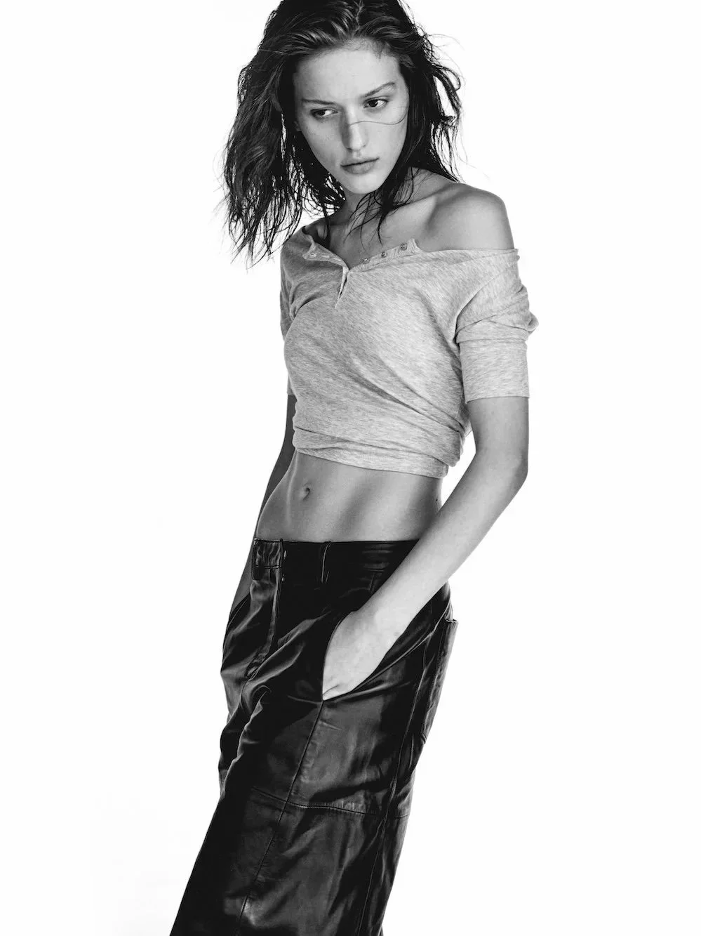 Black and white photo of a young woman with wavy hair, wearing a loose off-shoulder t-shirt and leather skirt, standing with one hand in her pocket and looking to the side.