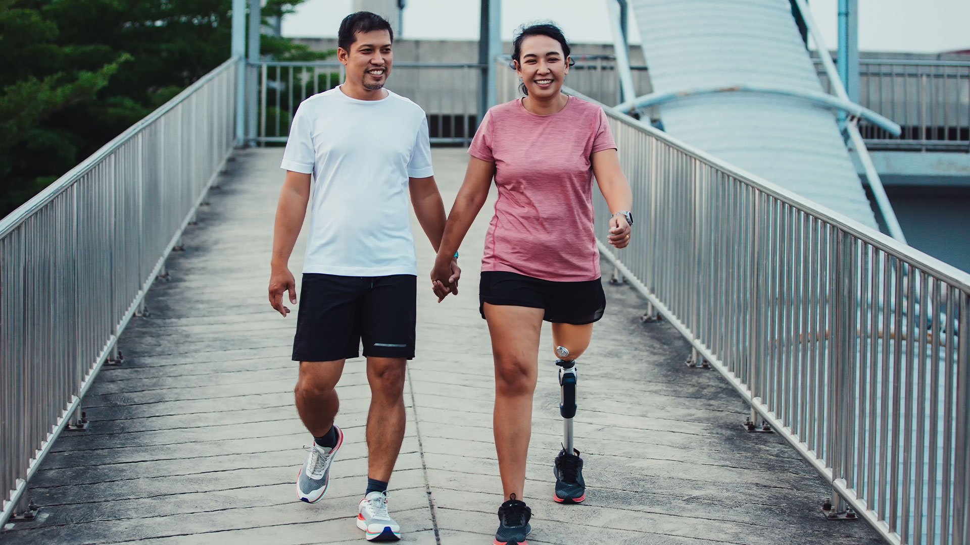 A smiling woman with a prosthetic leg walking hand in hand with a man on a bridge. Both are wearing athletic clothing, indicating they are on a casual walk or exercising together.