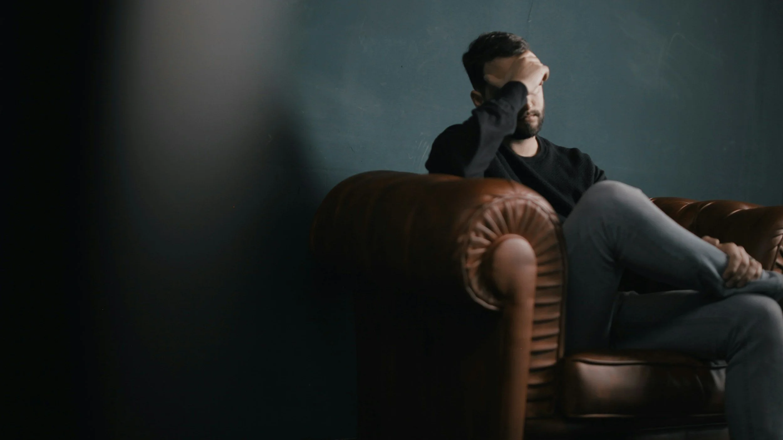 Man sitting on a leather couch, covering his face with his hand in a gesture of stress or fatigue, representing the emotional toll and struggles faced during difficult transitions.