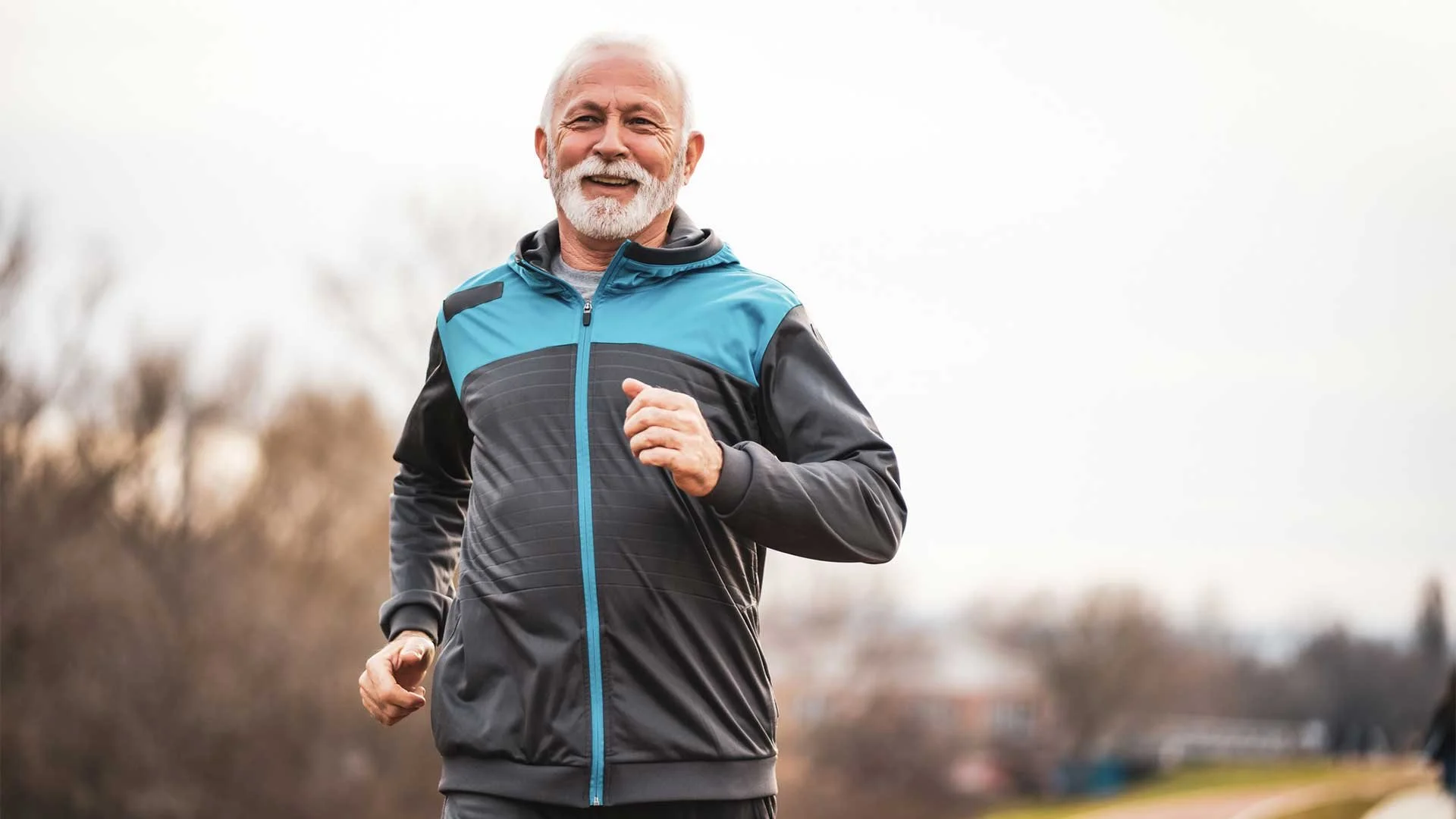 A senior man with a white beard is jogging outdoors while wearing a blue and gray jacket. He is smiling and appears to be enjoying his run, with a blurred background of trees and an overcast sky.
