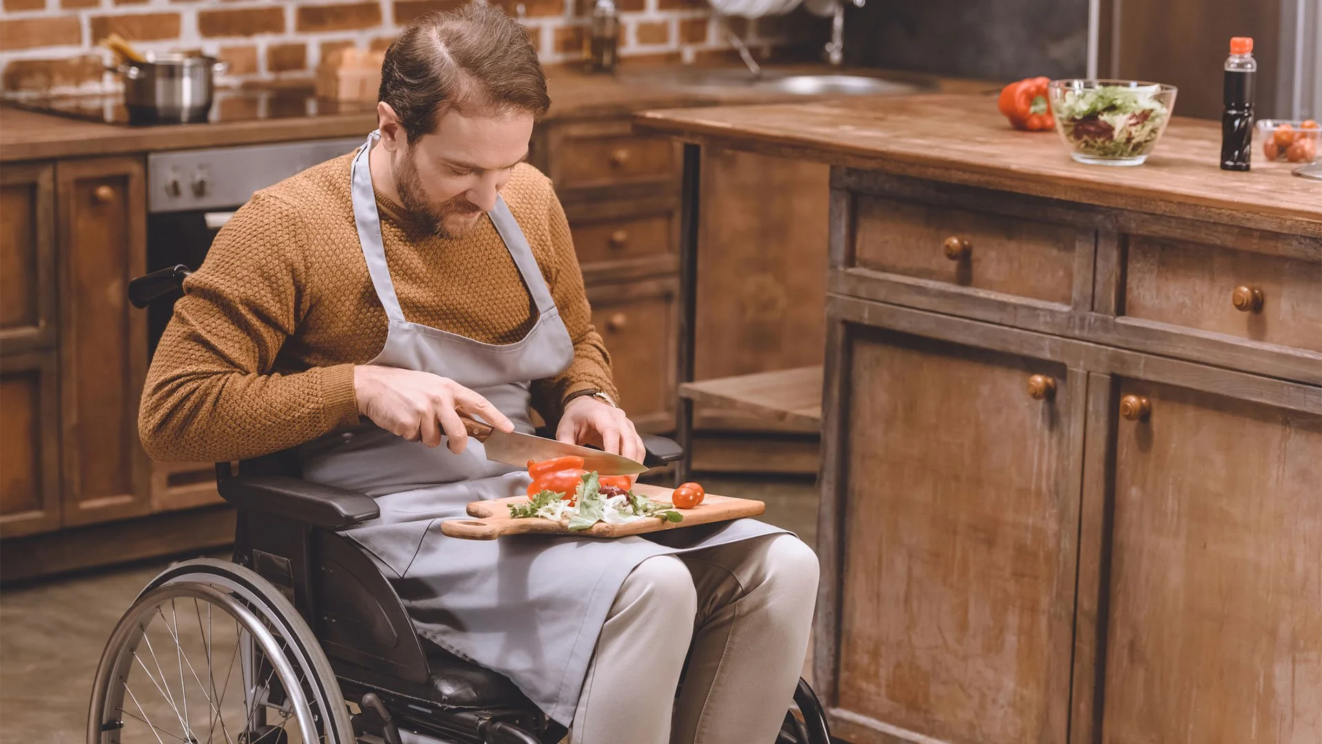 A man in a wheelchair is preparing a meal in a rustic kitchen. He is chopping vegetables on a cutting board placed on his lap, with various kitchen items and ingredients around him.