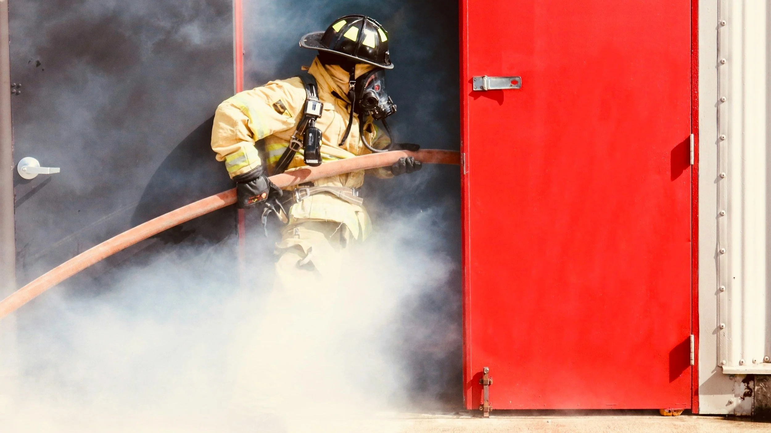 Firefighter in full gear emerging from a building with a red door, holding a hose amidst smoke, symbolizing the bravery and service of first responders.