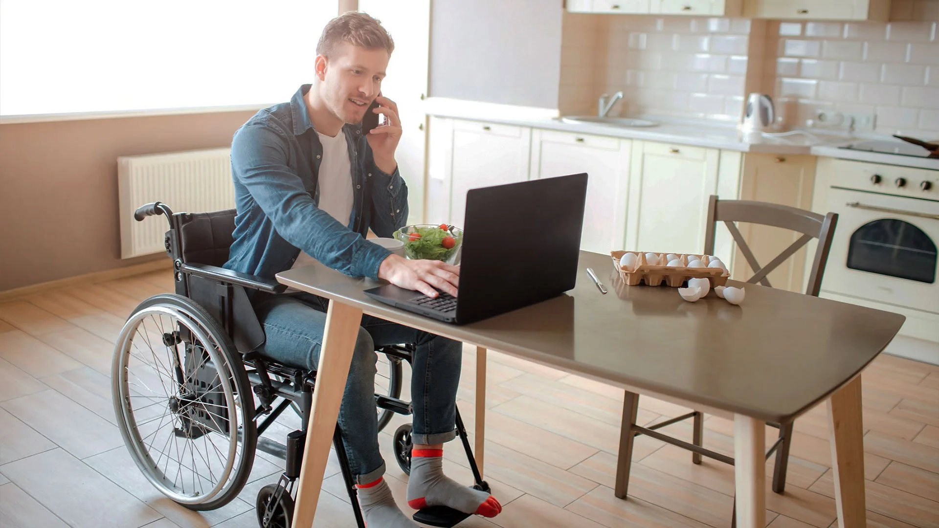 A man in a wheelchair is sitting at a kitchen table, talking on the phone while working on his laptop. He has a salad bowl next to him and the kitchen background is well-lit.
