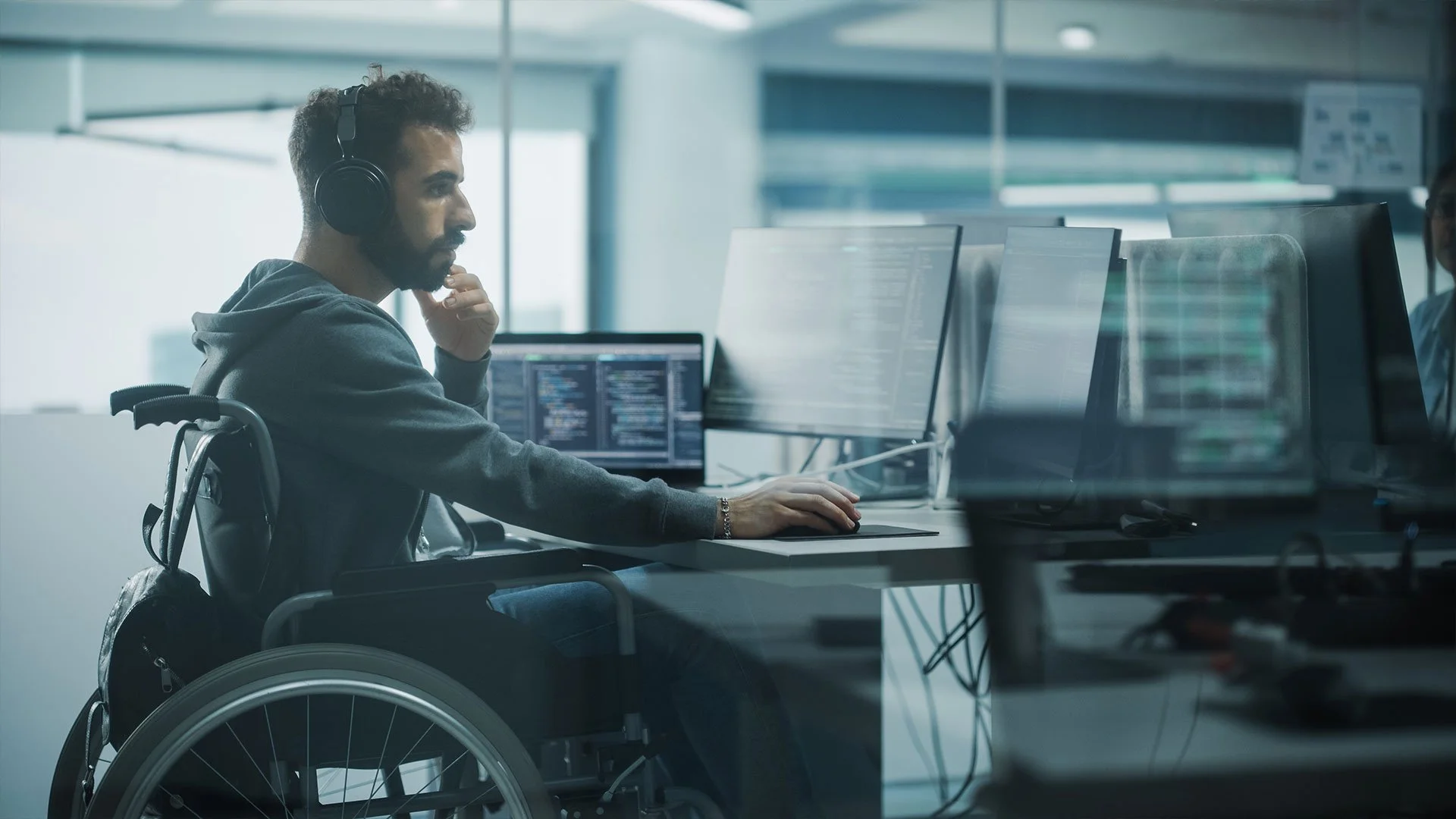A man in a wheelchair wearing headphones is focused on his work at a computer desk with multiple monitors in an office setting.