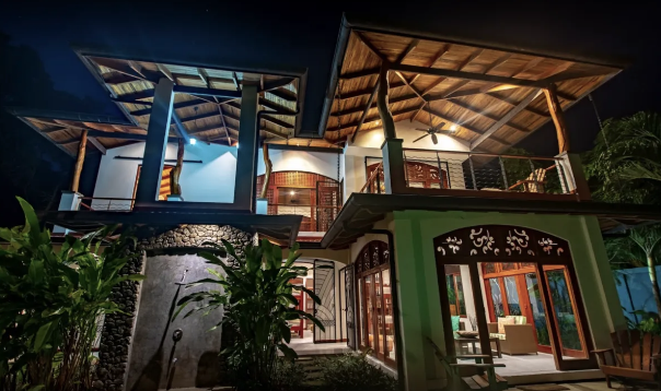 Outdoor dining area with wooden table and chairs, bar counter with stools, and windows with iron grills. The space has a tiled floor and wooden ceiling beams, creating a rustic and airy atmosphere.