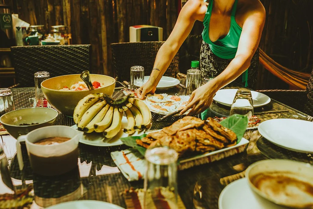 Person setting a dining table with bananas, fruit salad, and bread.