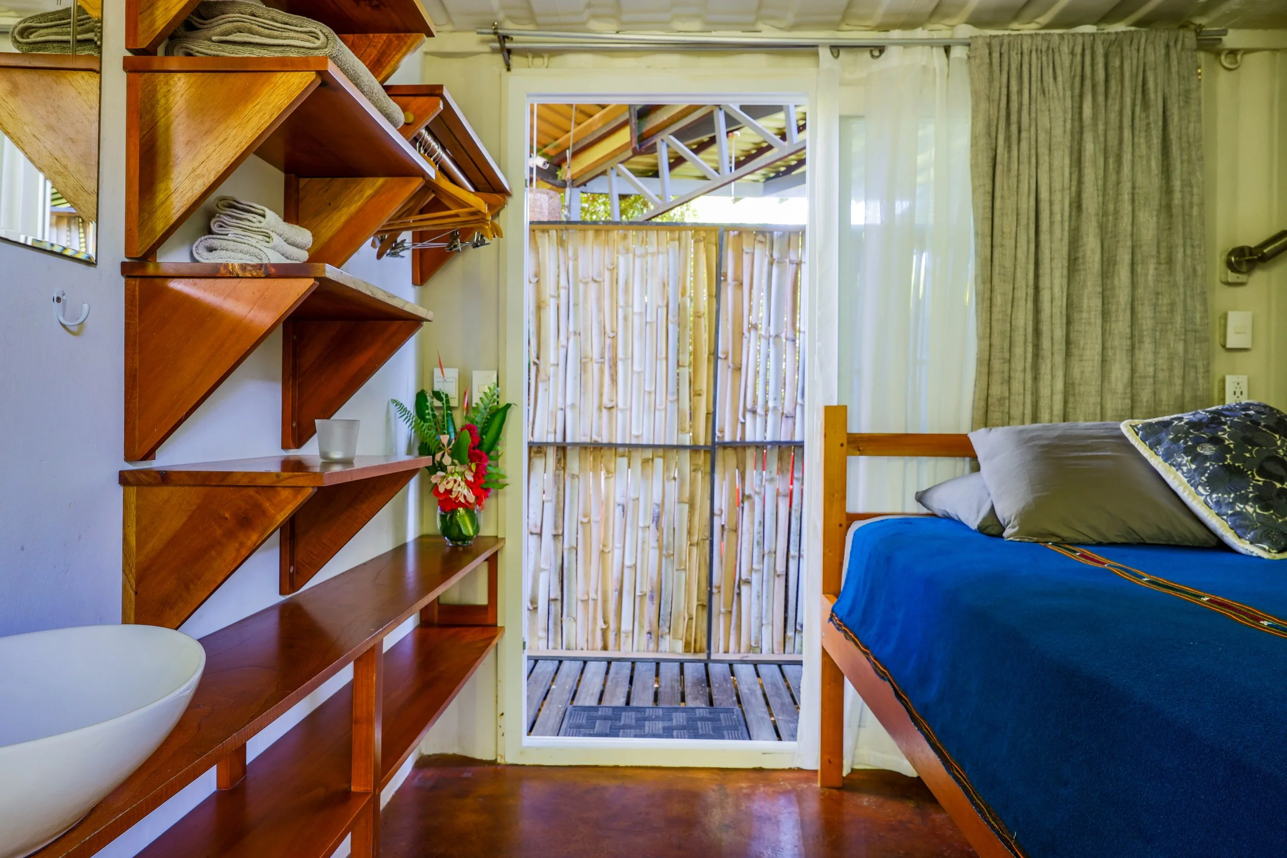 Interior of a small room featuring wooden shelves, folded towels, a blue-covered bed, and a door leading to a bamboo-fenced area. Decorative plants and curtains are also visible.