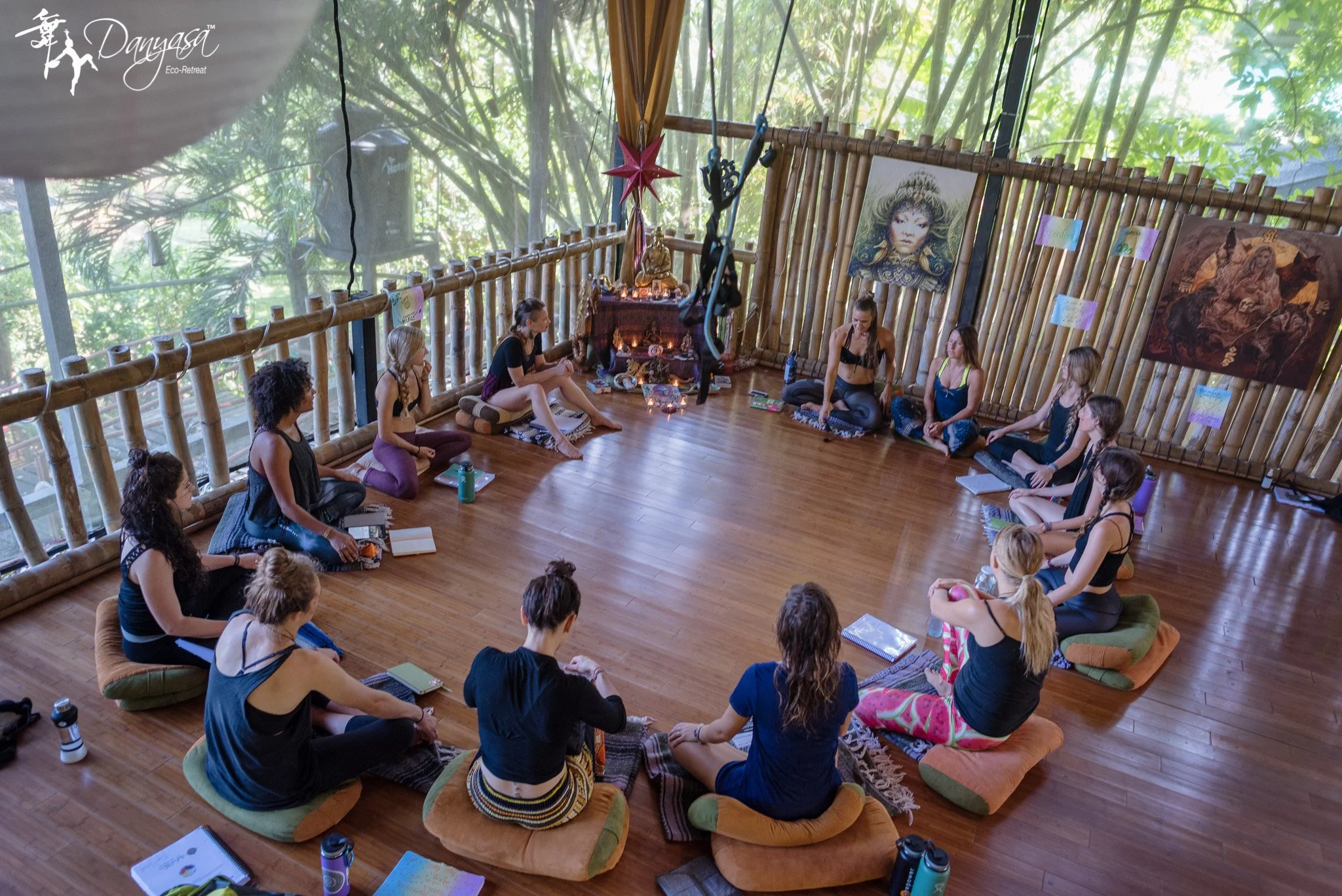 Group of people sitting in a circle on cushions in a bamboo eco-retreat space, engaging in a workshop or meditation session.