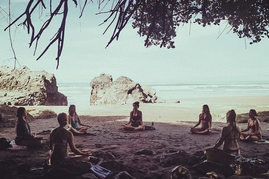 A group of women practicing yoga on a beach under a canopy of trees, with large rocks and ocean waves in the background.
