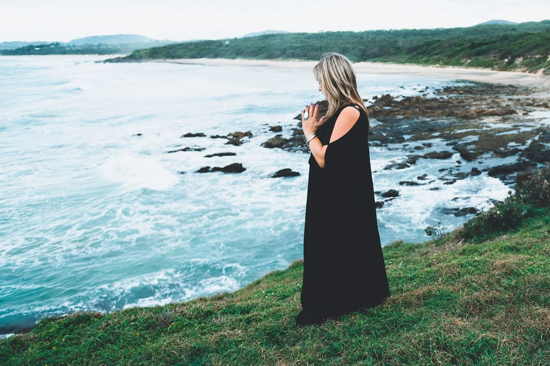 Woman in black dress standing on grassy cliff near the ocean, with her hands in prayer position and her eyes closed, facing the water.