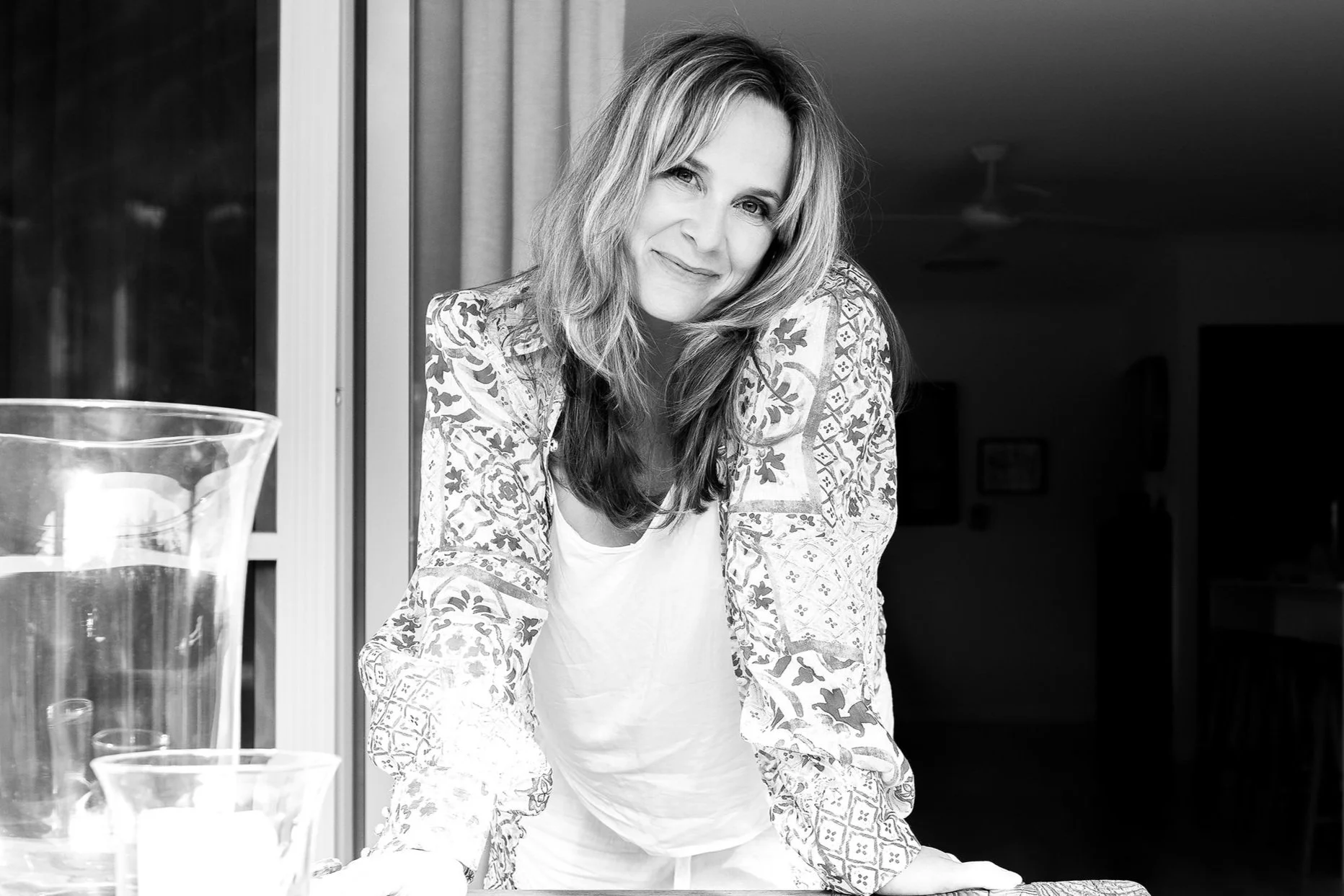 A woman with shoulder-length hair smiling and leaning forward on a table in a room.
