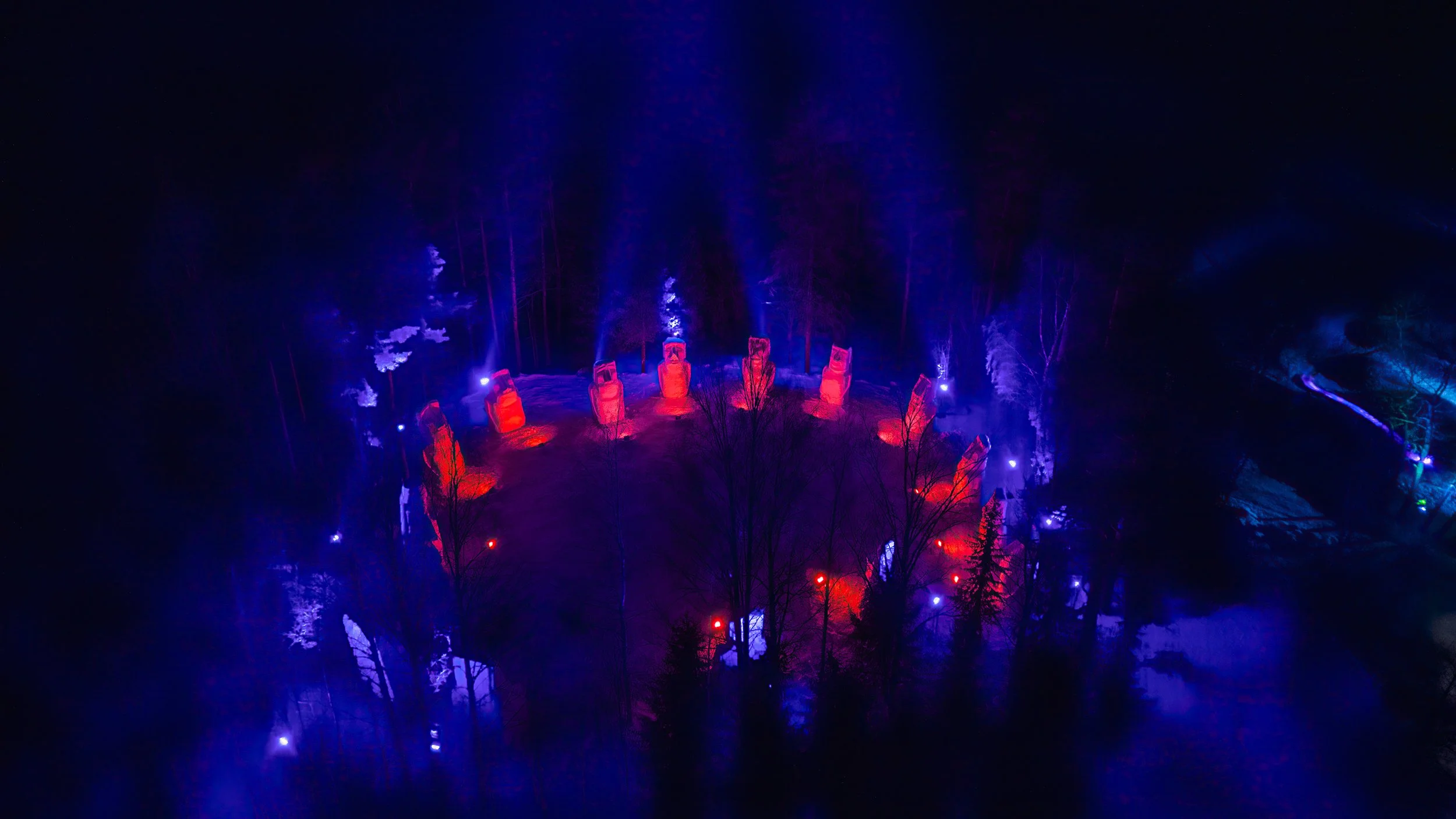 Aerial night view of a circular formation of stone statues illuminated with red and purple lights, surrounded by trees with some lighting effects in a dark forested area.