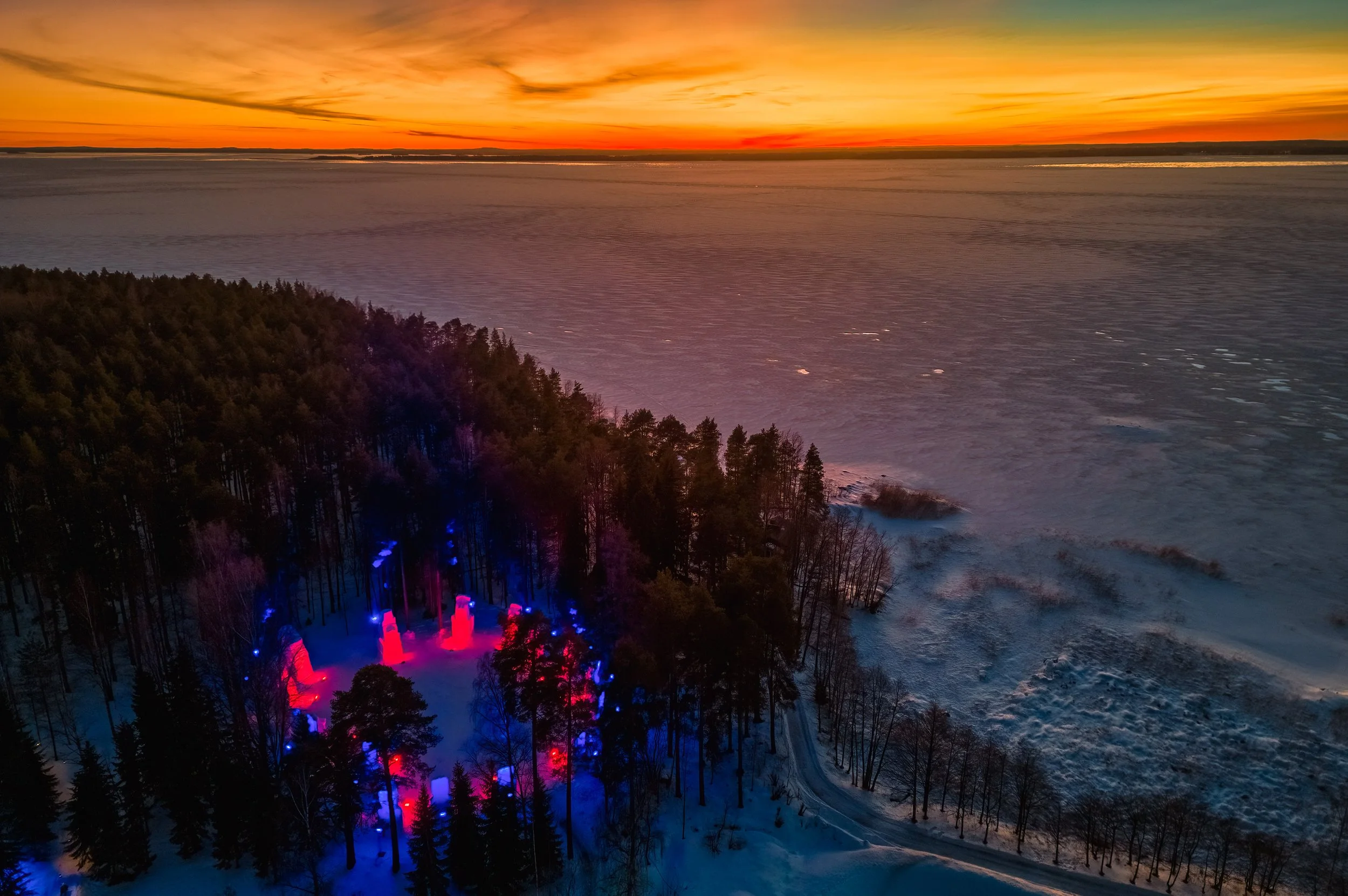 Aerial view of a snowy forest next to a large body of water during sunset, with colorful lights illuminating the trees.