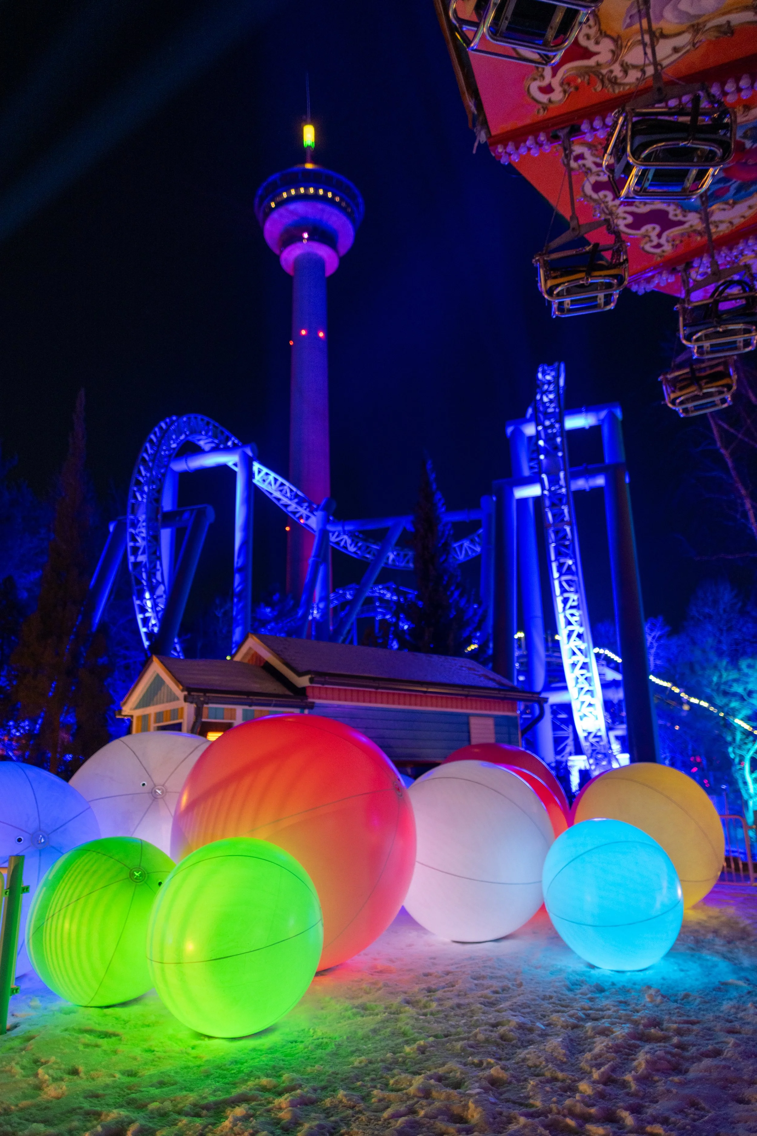 Night scene of a colorful amusement park with illuminated glowing spheres in the foreground and a roller coaster and observation tower in the background.