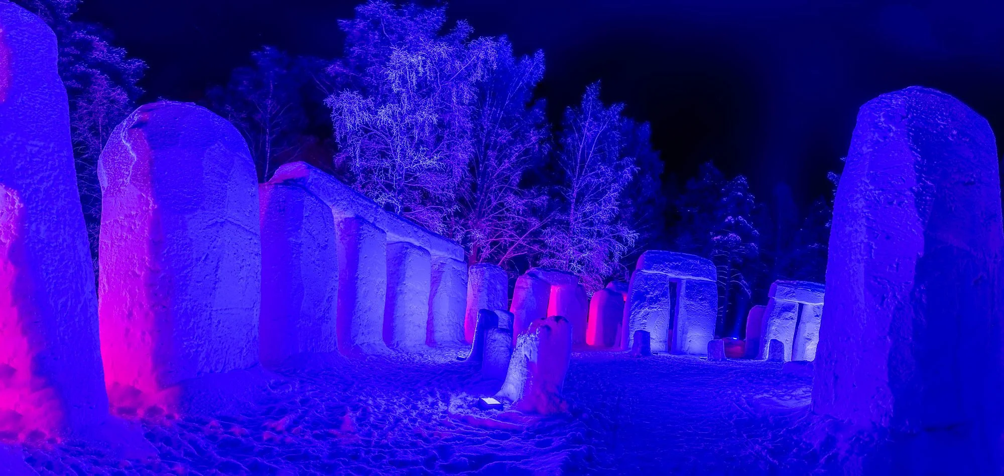 Ice sculptures illuminated with colorful purple, blue, and pink lights at night. Snow-covered ground with leafless trees in the background.