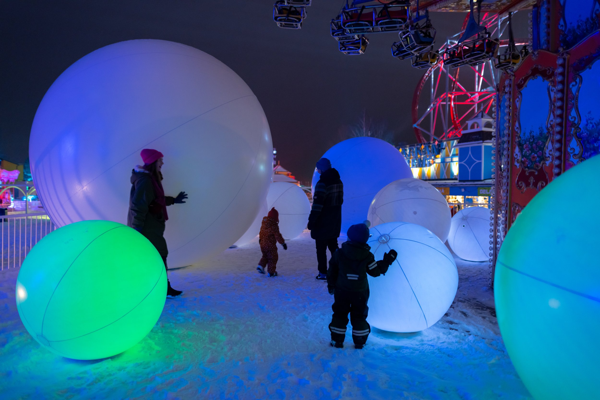 People walking among glowing, colorful, illuminated spheres at an amusement park during nighttime.