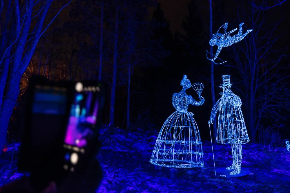 Blue wireframe illuminated sculptures of a woman in a dress, a man in a suit and top hat, and a flying creature holding a bow and arrow, set against a dark outdoor background with trees.