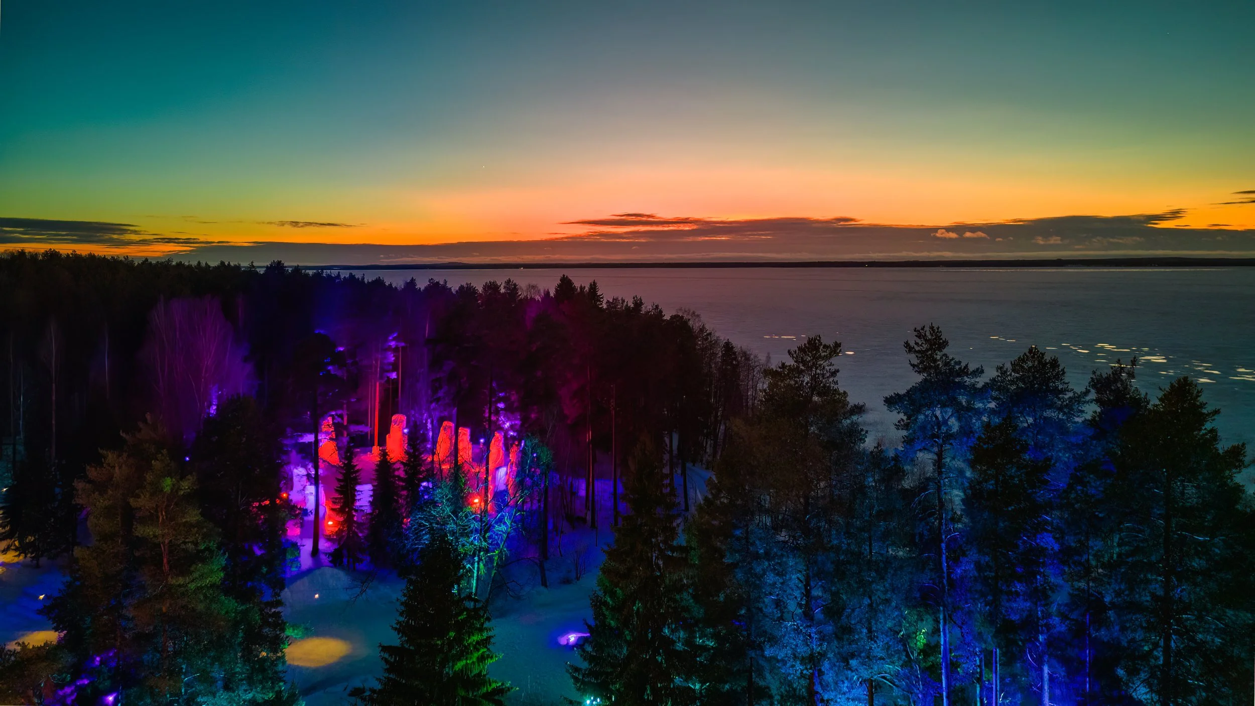 Nighttime view of a forest with colorful rainbow lights illuminating trees, overlooking a body of water during sunset.