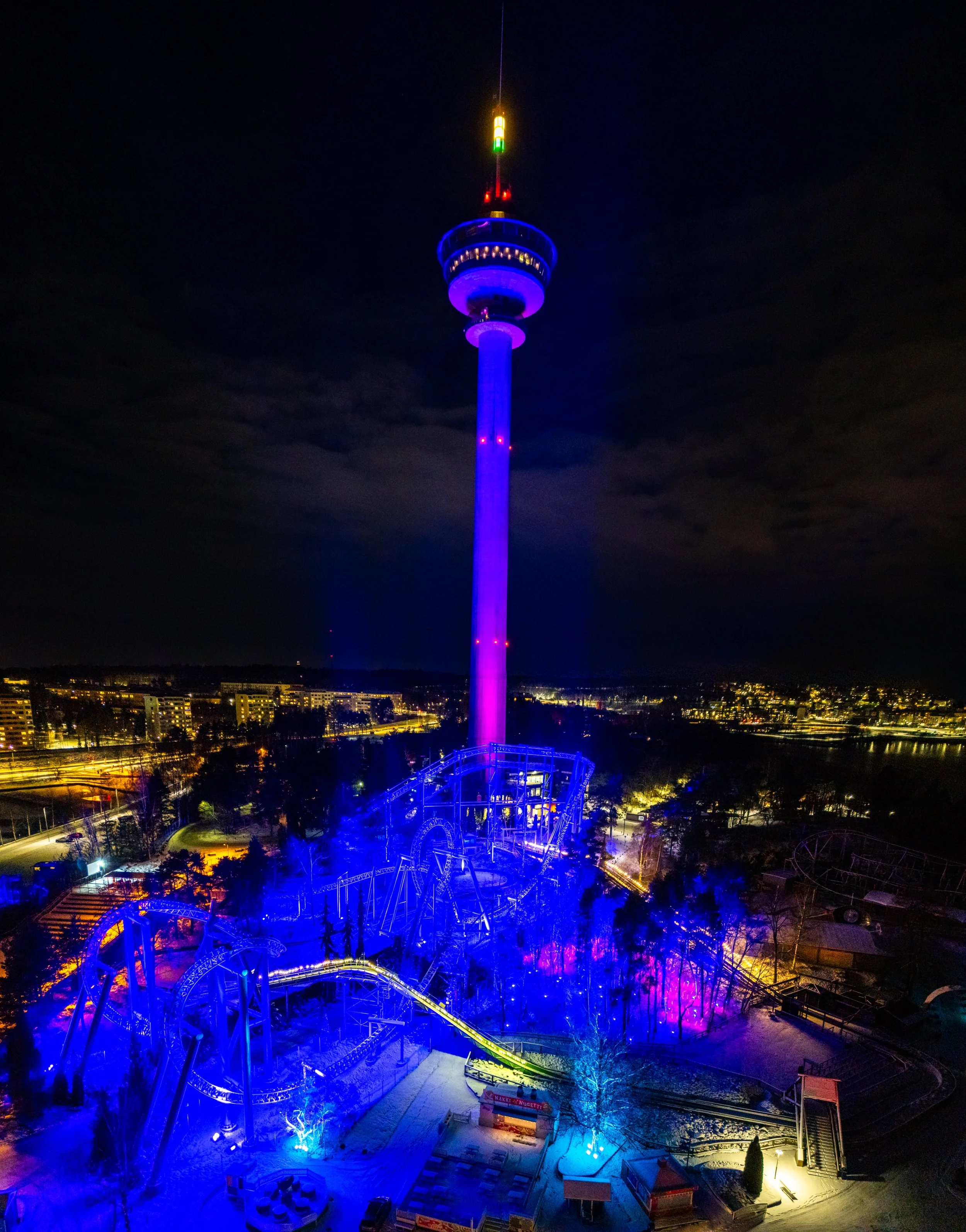 Nighttime view of a tall tower and an amusement park with roller coasters illuminated in blue and purple lights.