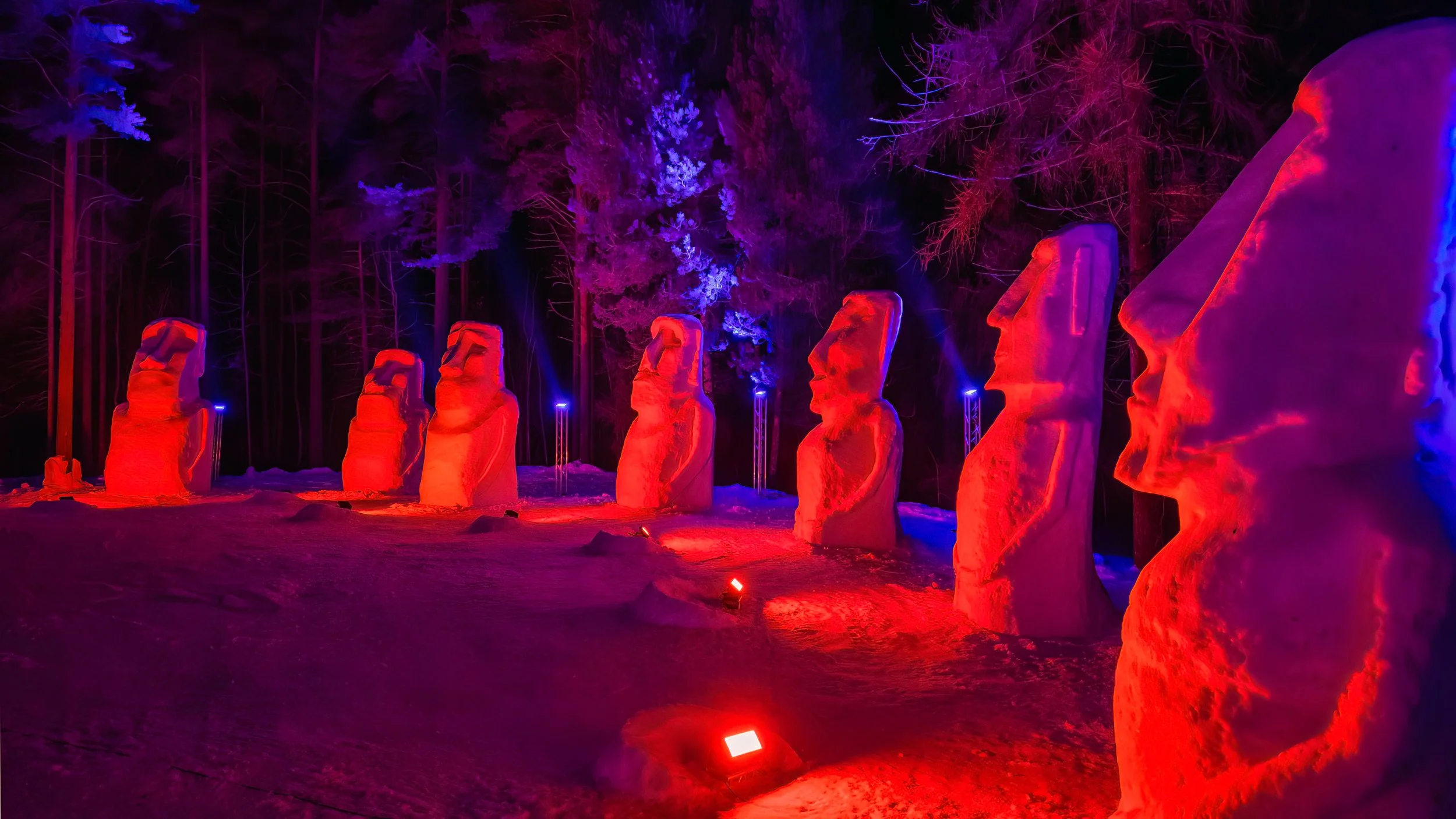 Row of illuminated Moai statues during nighttime with colorful lights in a snowy setting.