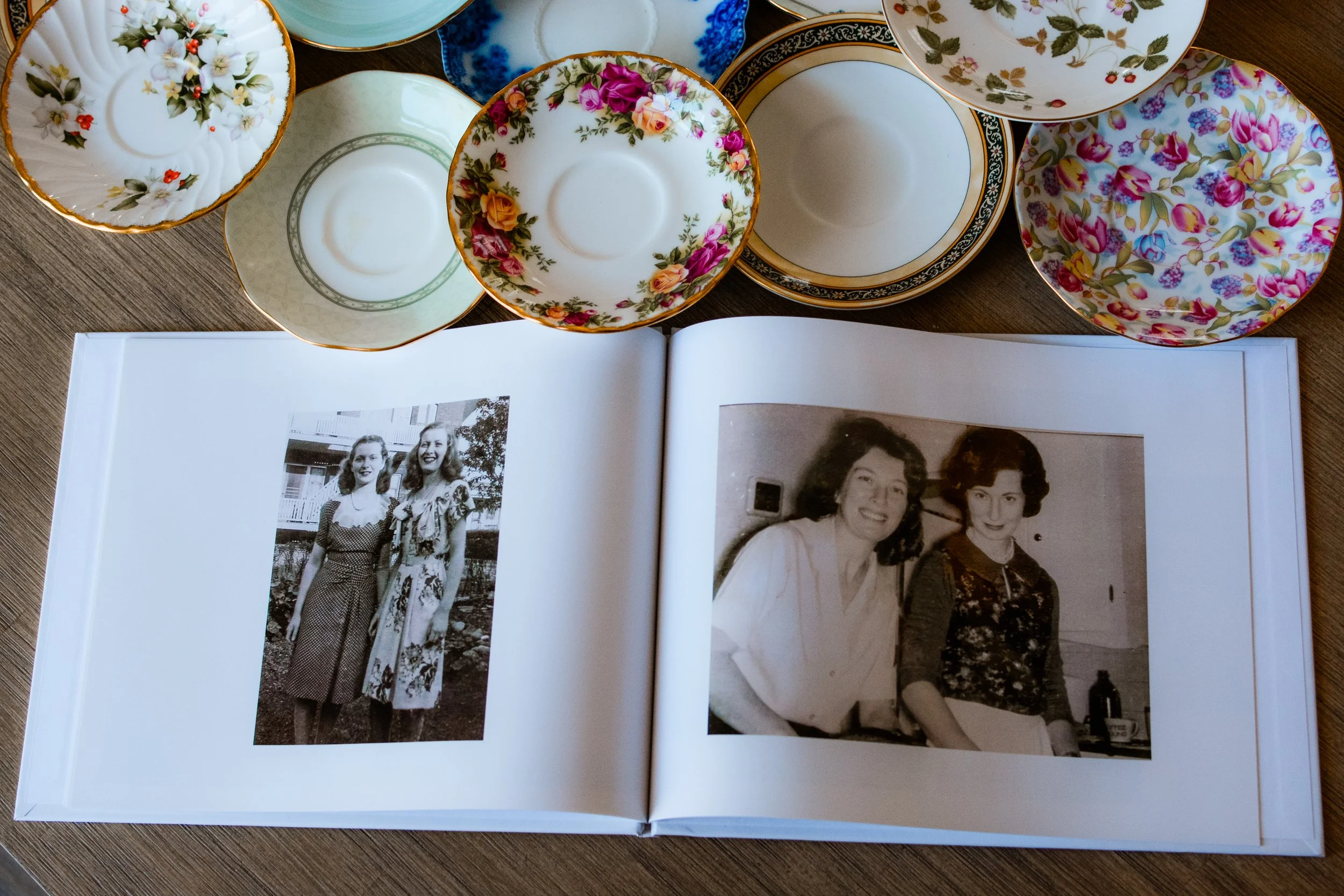 A photo album lies open on the table surrounded by an assortment of colorful china plates. The album lies open to two black and white images, each of two young women, smiling.