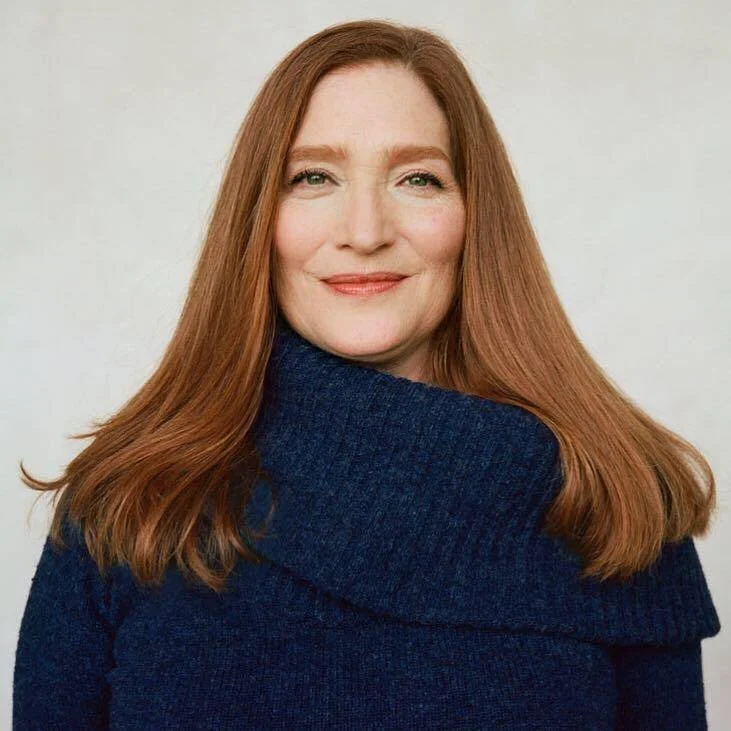 Portrait of a woman with long red hair wearing a dark blue sweater, smiling at the camera against a plain light background.