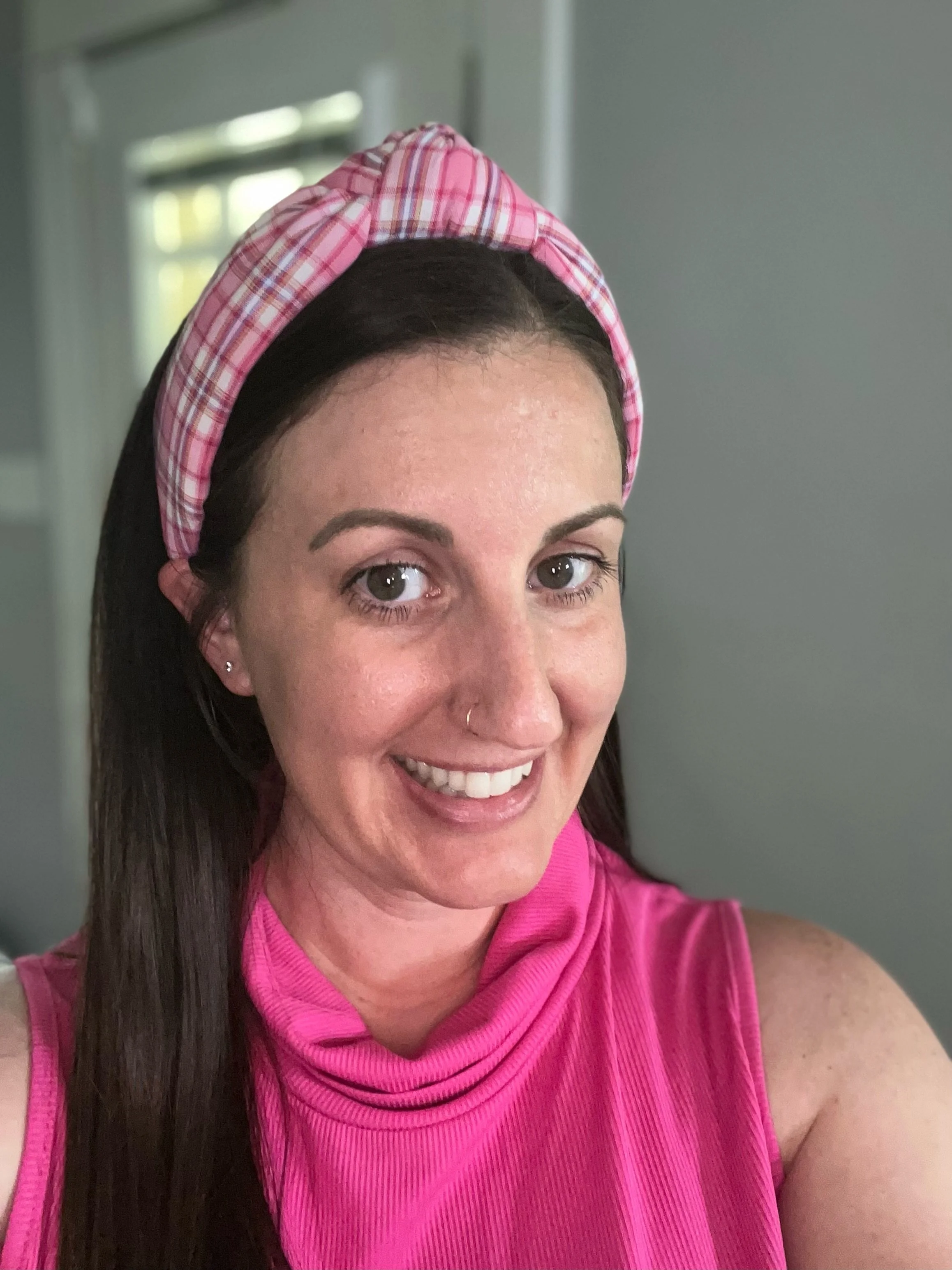 A woman with long dark hair wearing a pink sleeveless top and pink plaid headband smiling in front of a neutral background.