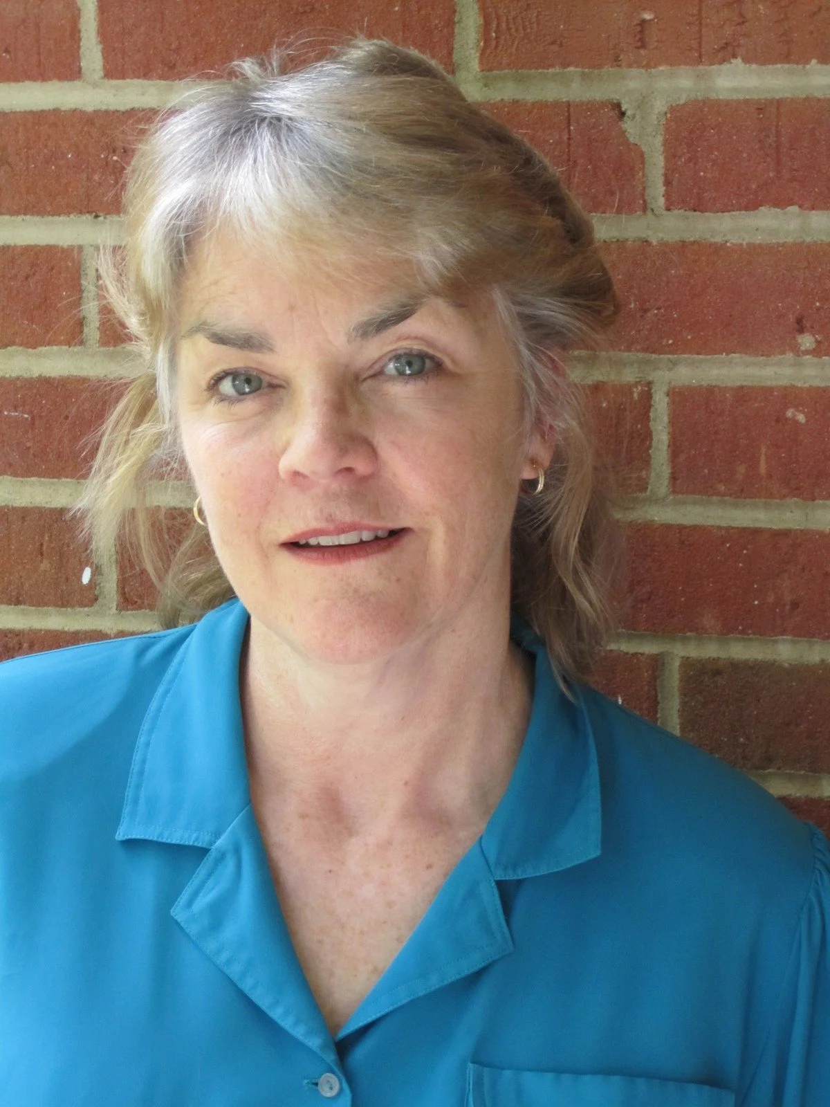 A woman with light skin, shoulder-length blonde hair, green eyes, wearing a blue collared shirt and small hoop earrings, standing against a brick wall.