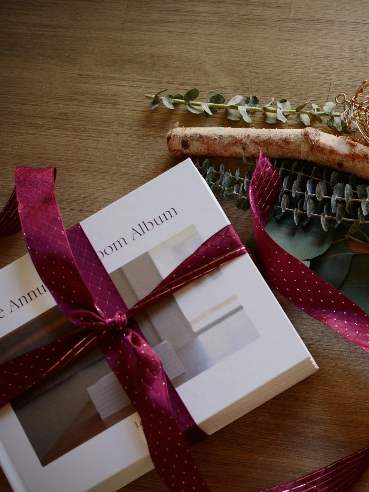 A photo of a wedding album tied with a burgundy ribbon, resting on a wooden surface next to eucalyptus leaves and a piece of driftwood.